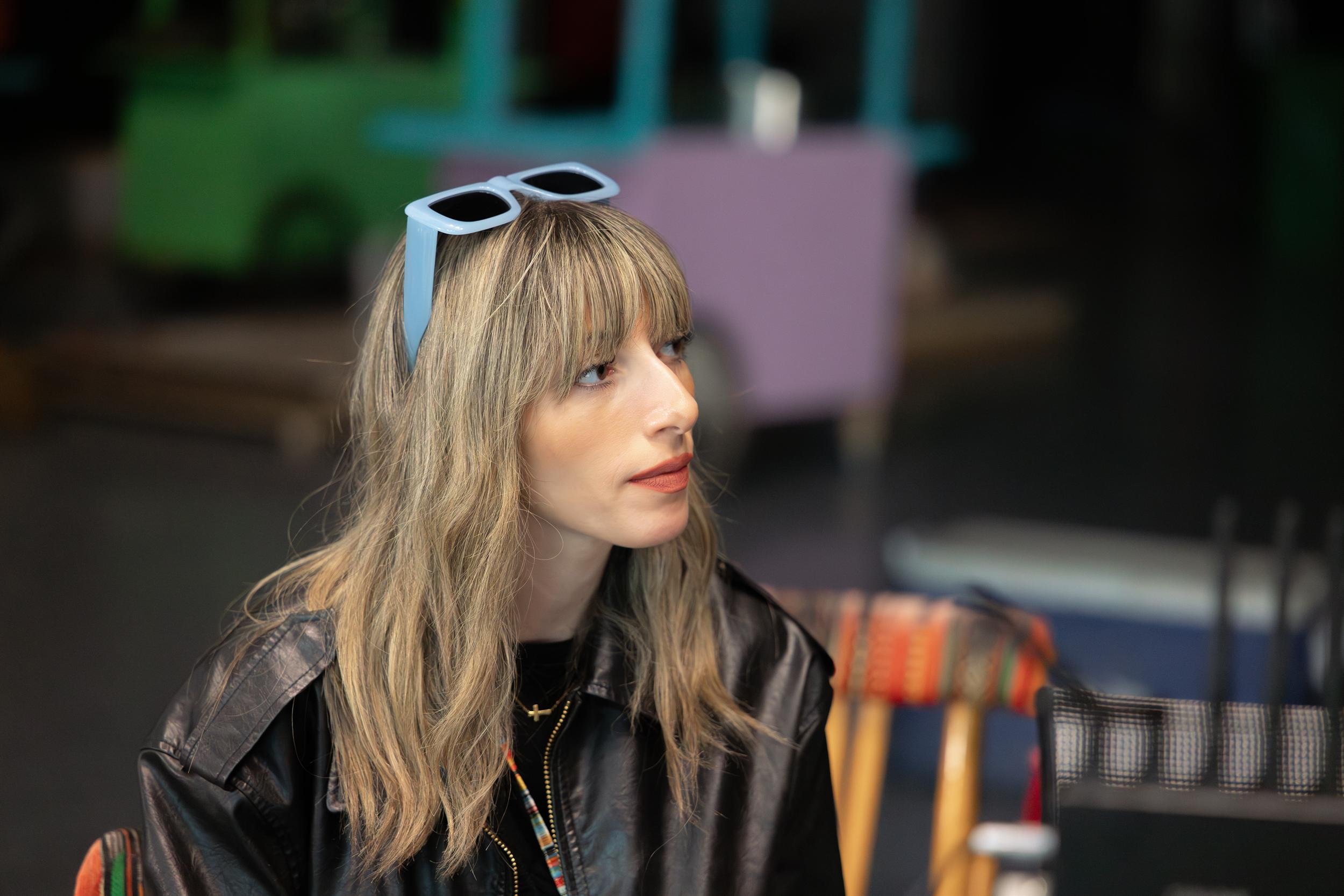 Ahlan Simsim unit still: a woman in a black leather jacket sits on set before colorful wooden stands.