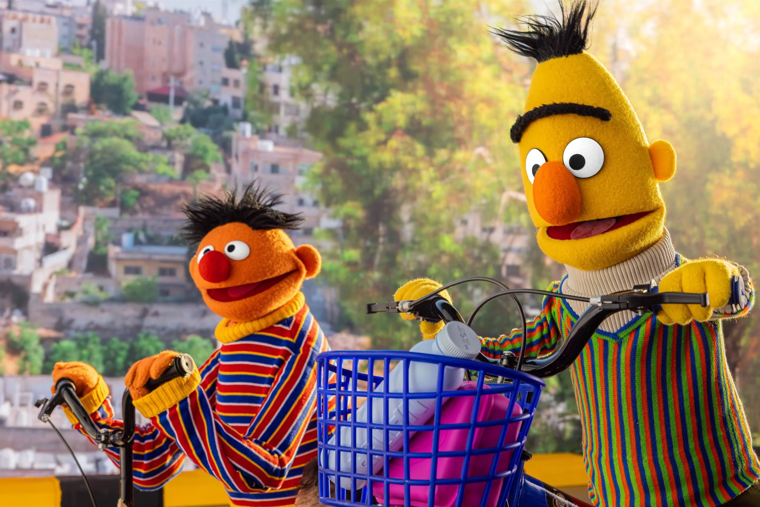 Ahlan Simsim key art: Ernie and Bert ride bicycles in front of a sunlit hillside town.