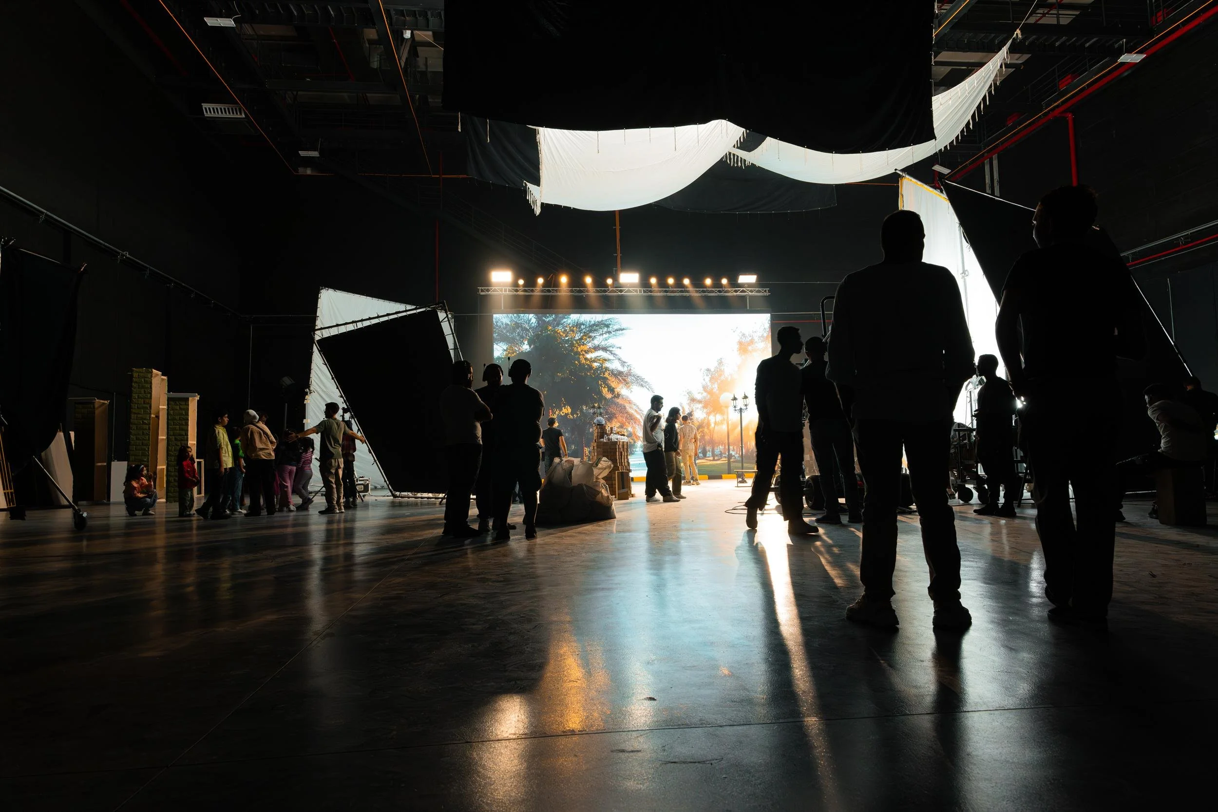 Ahlan Simsim unit still: a wide studio view of an LED park backdrop with crew and children near green brick pillars.