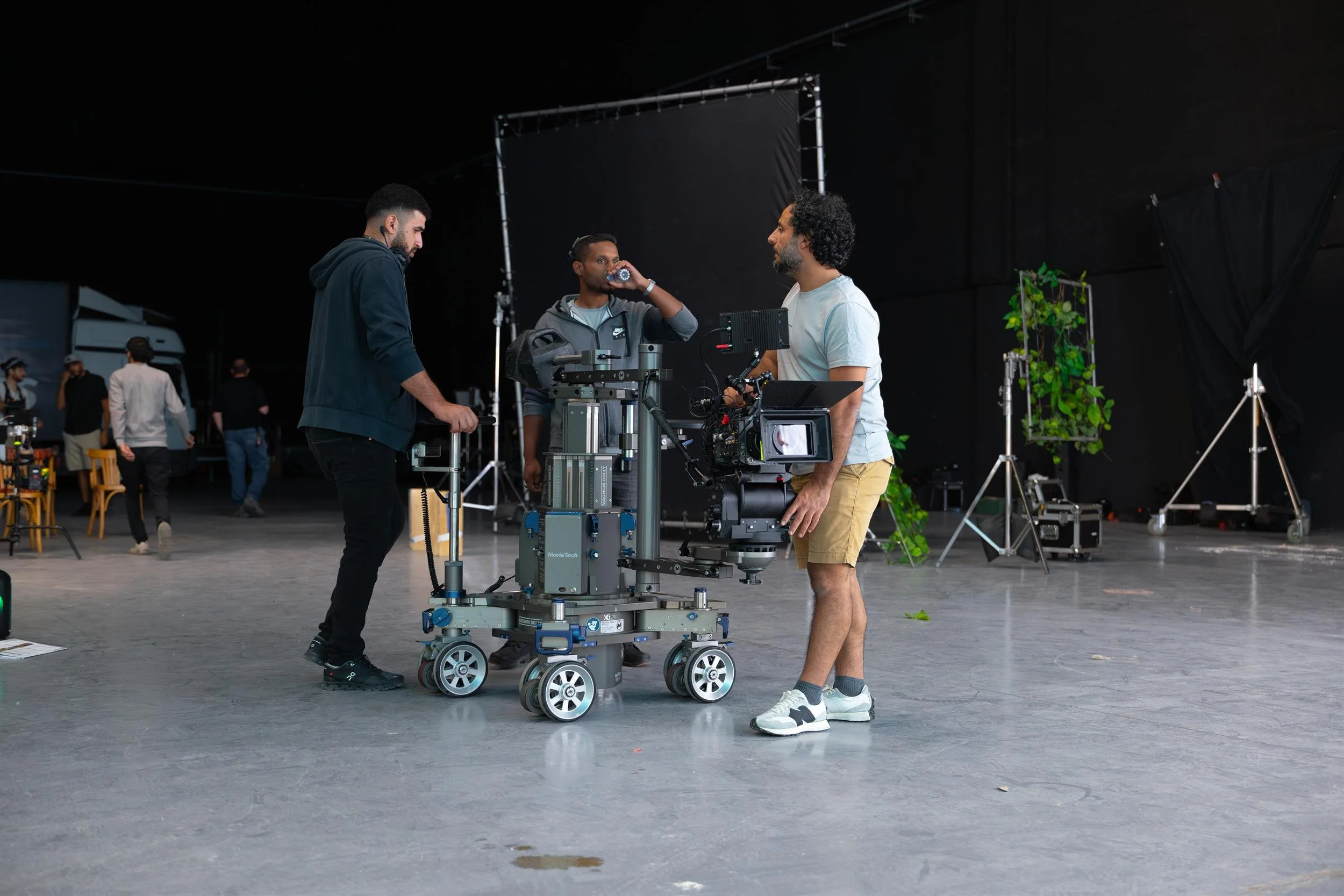 Ahlan Simsim unit still: crew lean over a camera on a wheeled dolly in a dark studio.