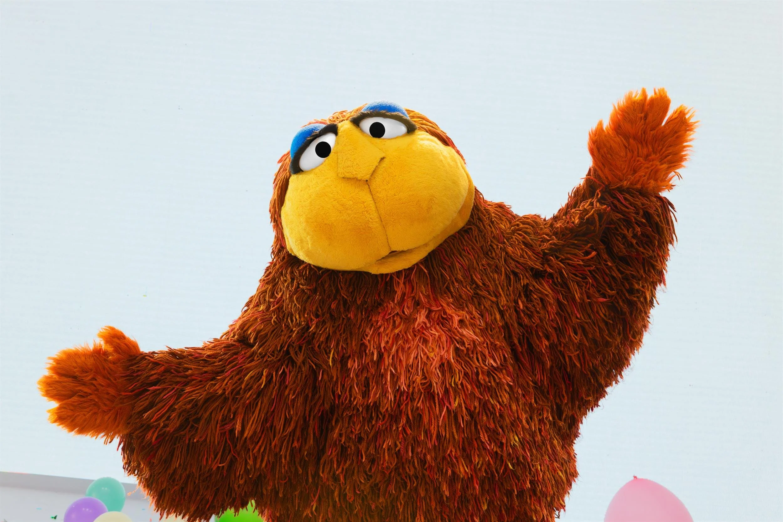 Ahlan Simsim key art: A large shaggy brown Muppet with a yellow face waves beside colorful balloons.