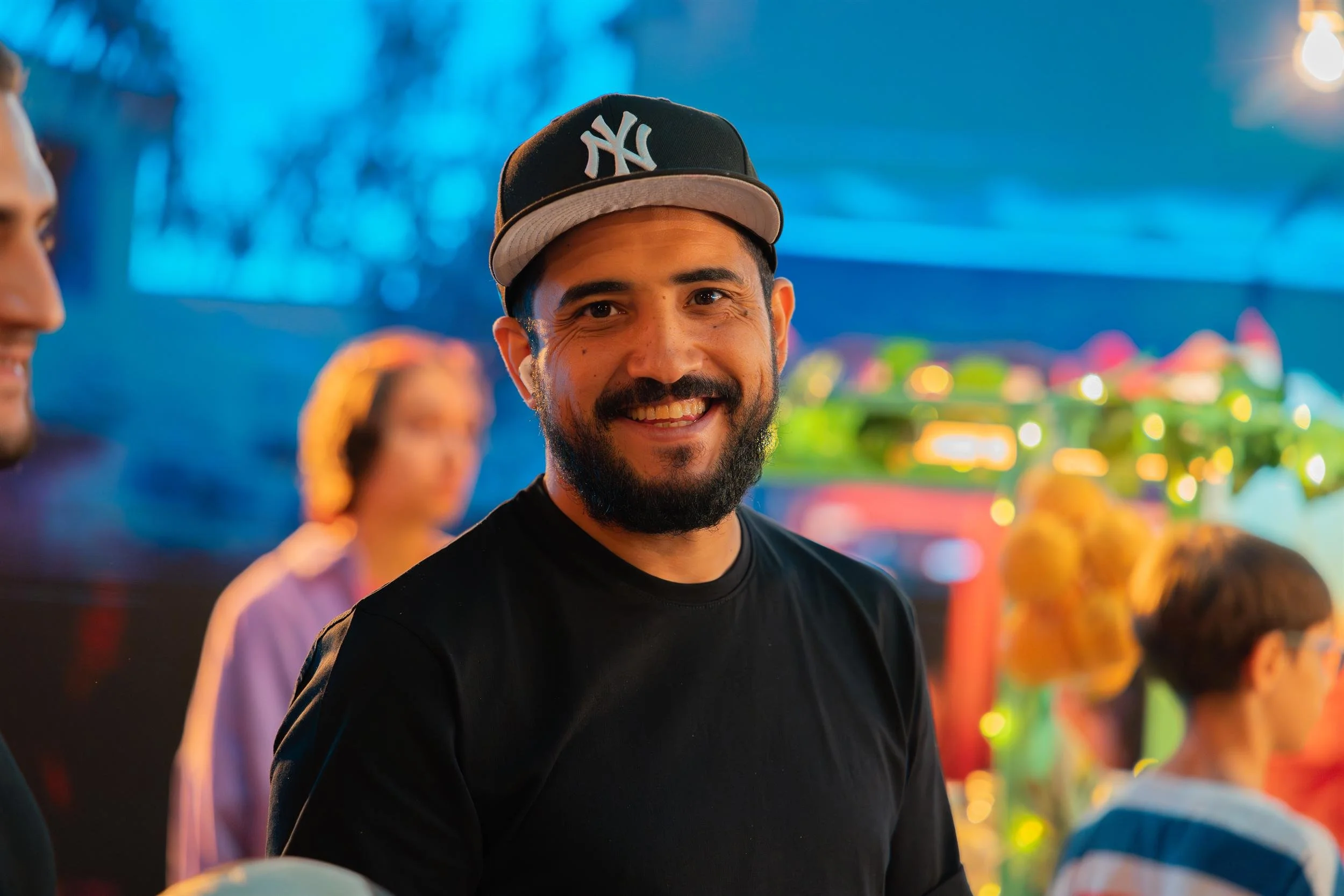 Ahlan Simsim unit still: a smiling crew member in a baseball cap on a night-market set.