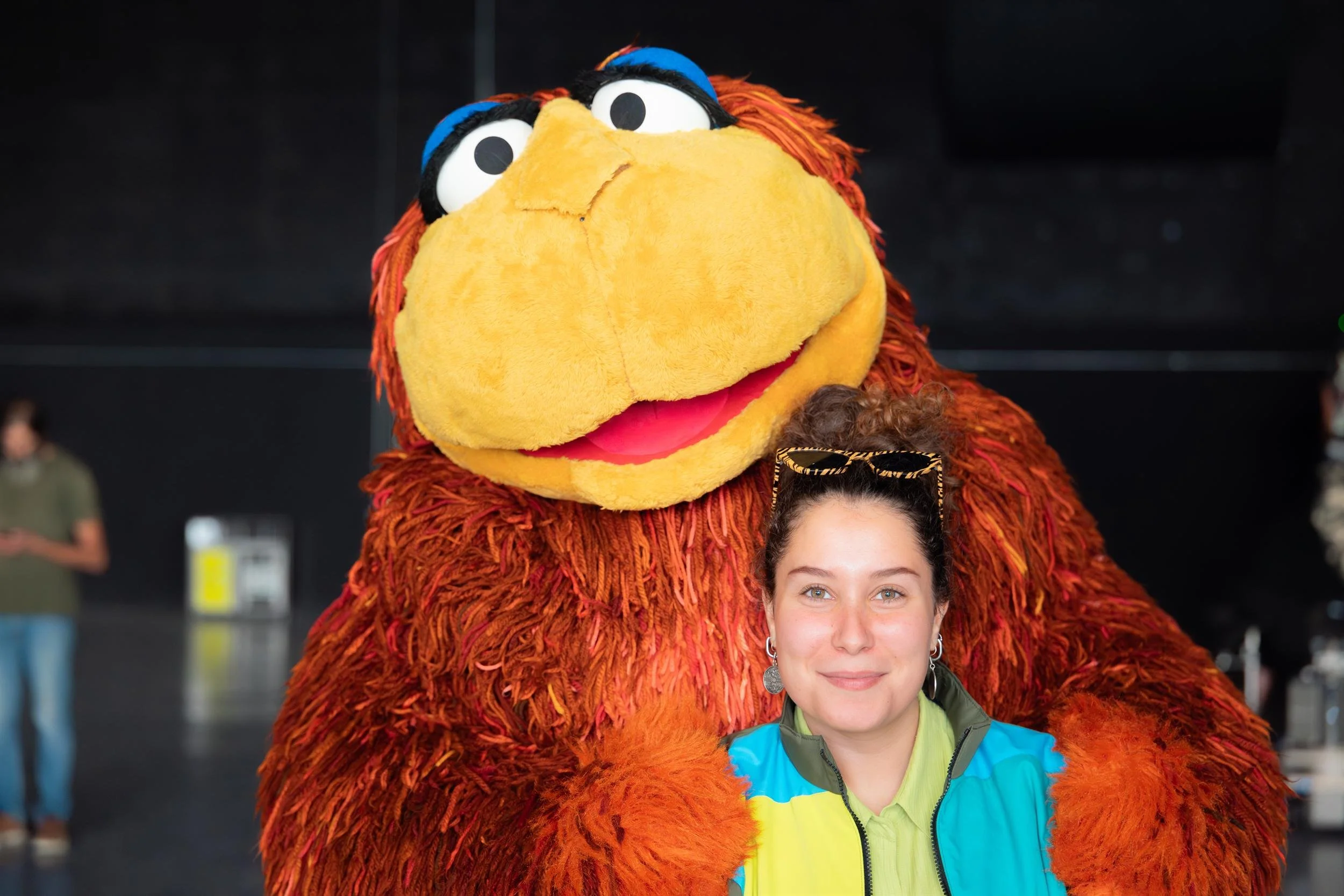 Ahlan Simsim unit still: a smiling woman is hugged by a large shaggy brown Muppet in a studio.