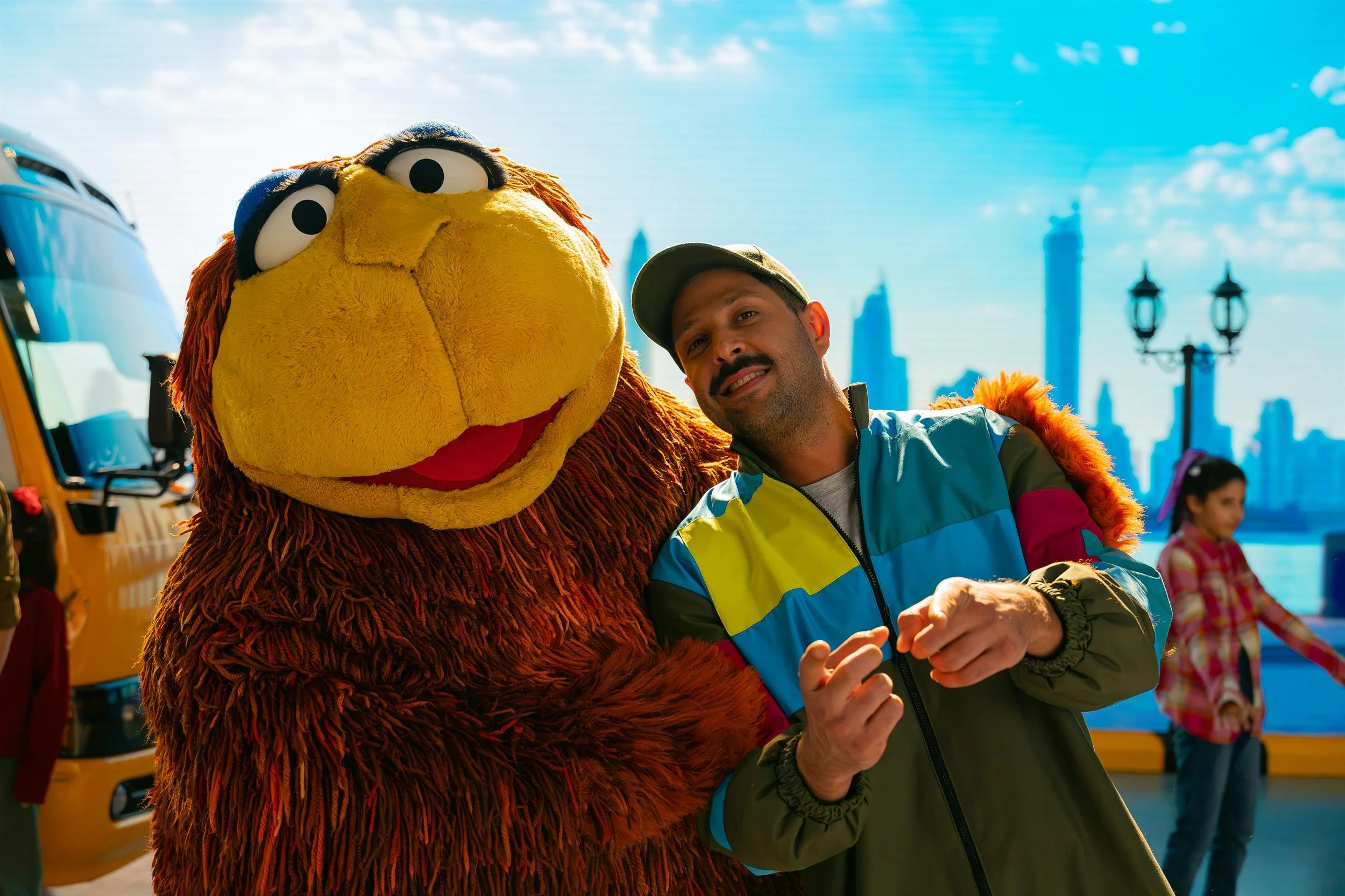 Ahlan Simsim unit still: a man in a color-block jacket points at the camera beside a large brown Muppet.
