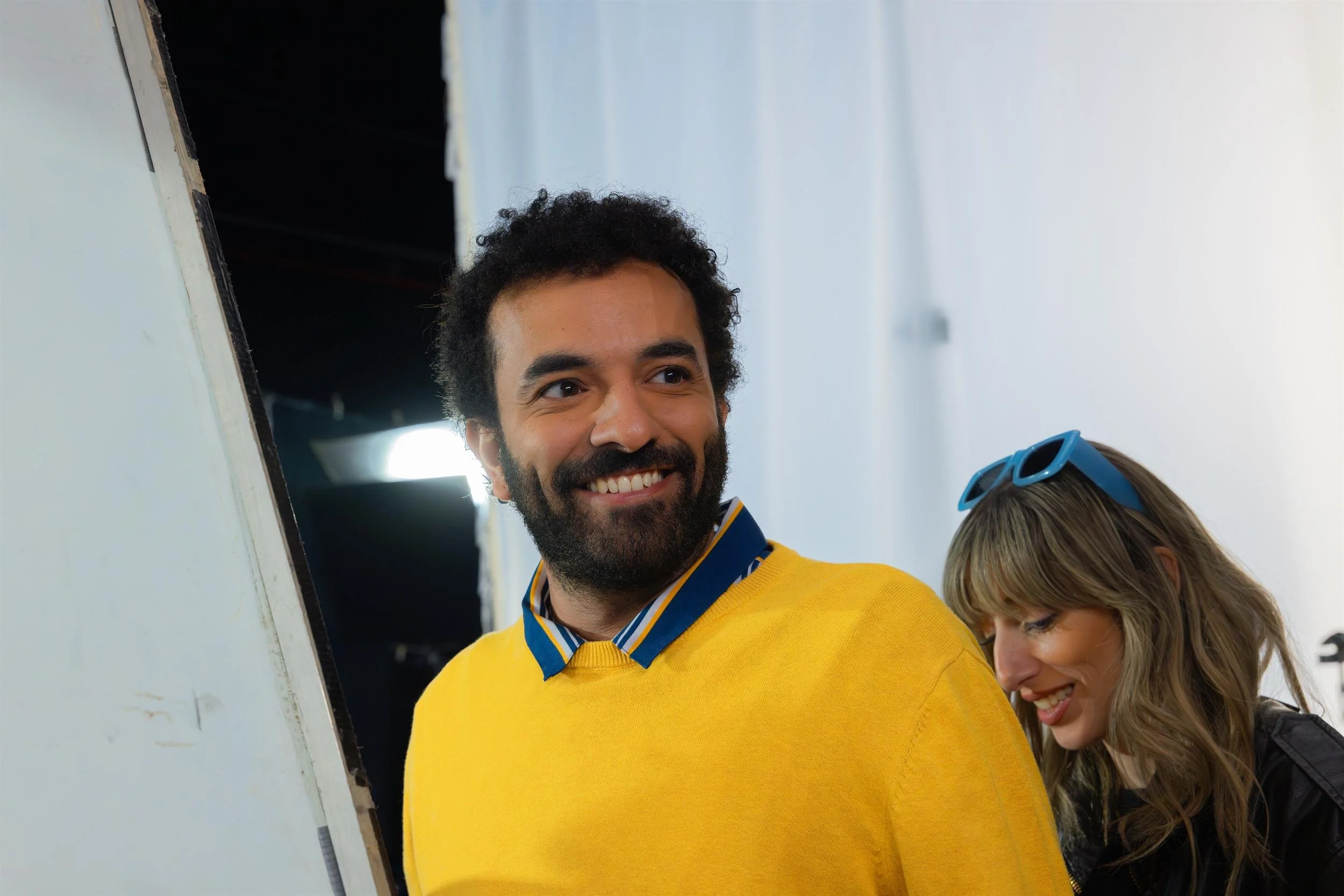 Ahlan Simsim unit still: a smiling man in a yellow sweater stands with a colleague before a white studio backdrop.