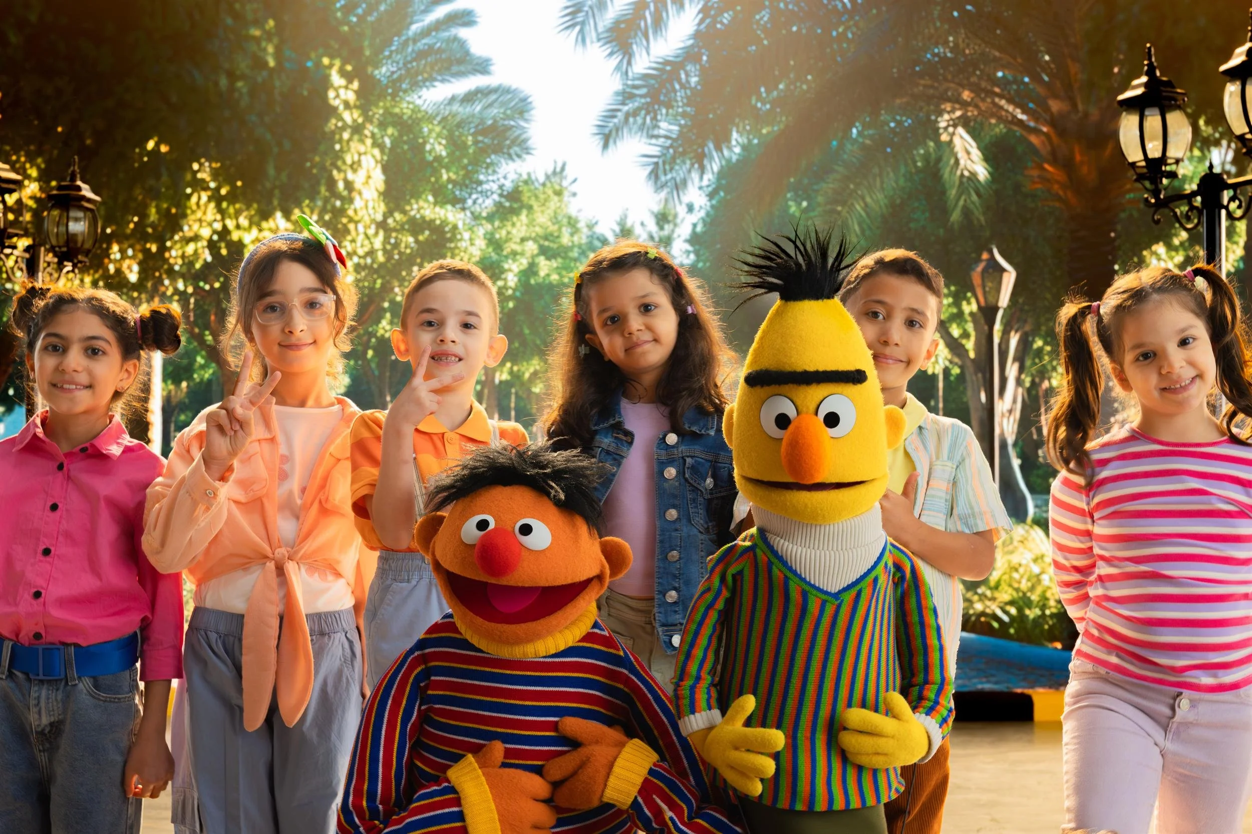 Ahlan Simsim unit still: Ernie, Bert, and six smiling children pose together in a sunlit park with palm trees.