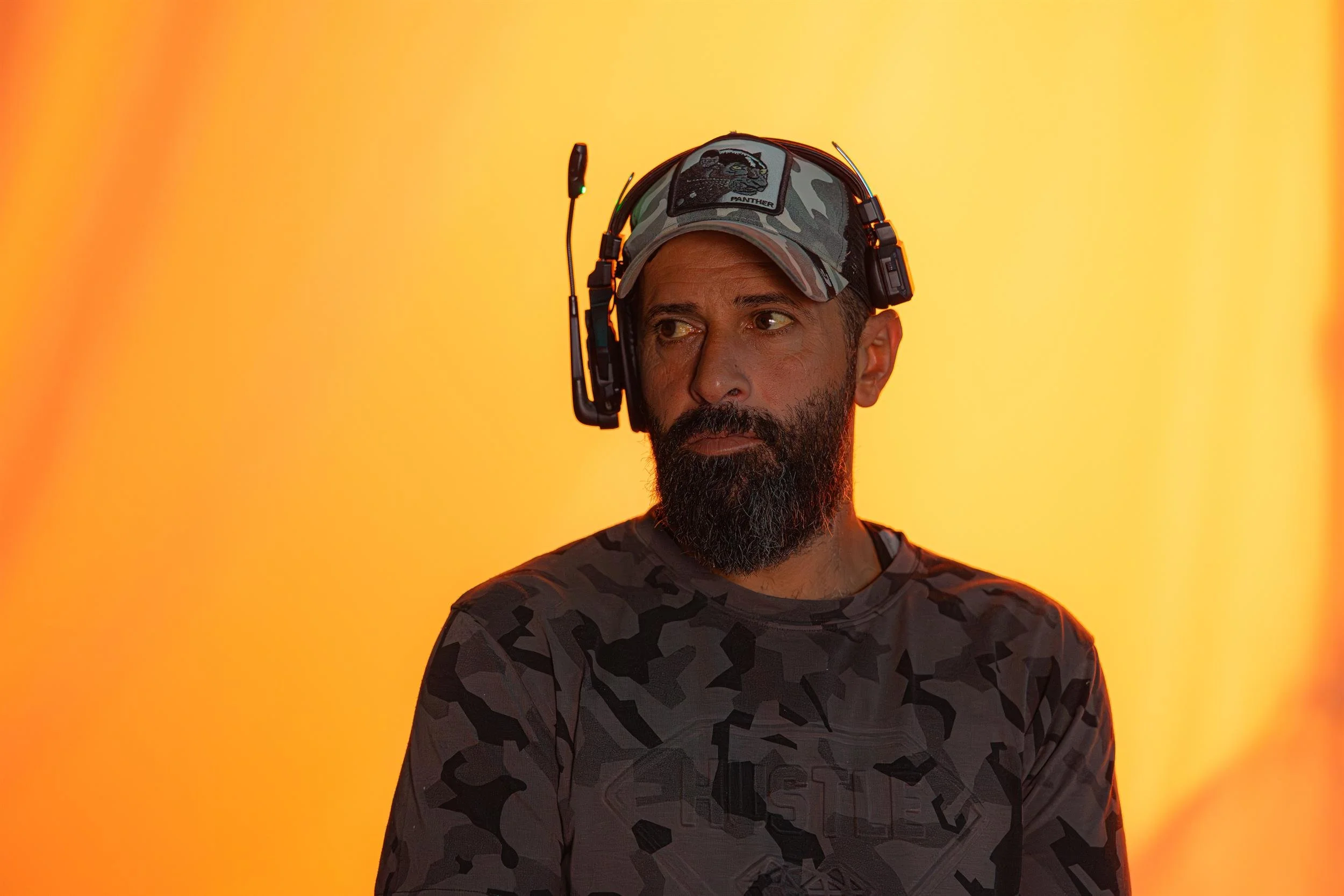 Ahlan Simsim unit still: a bearded crew member in a headset and camouflage hat against a bright orange backdrop.