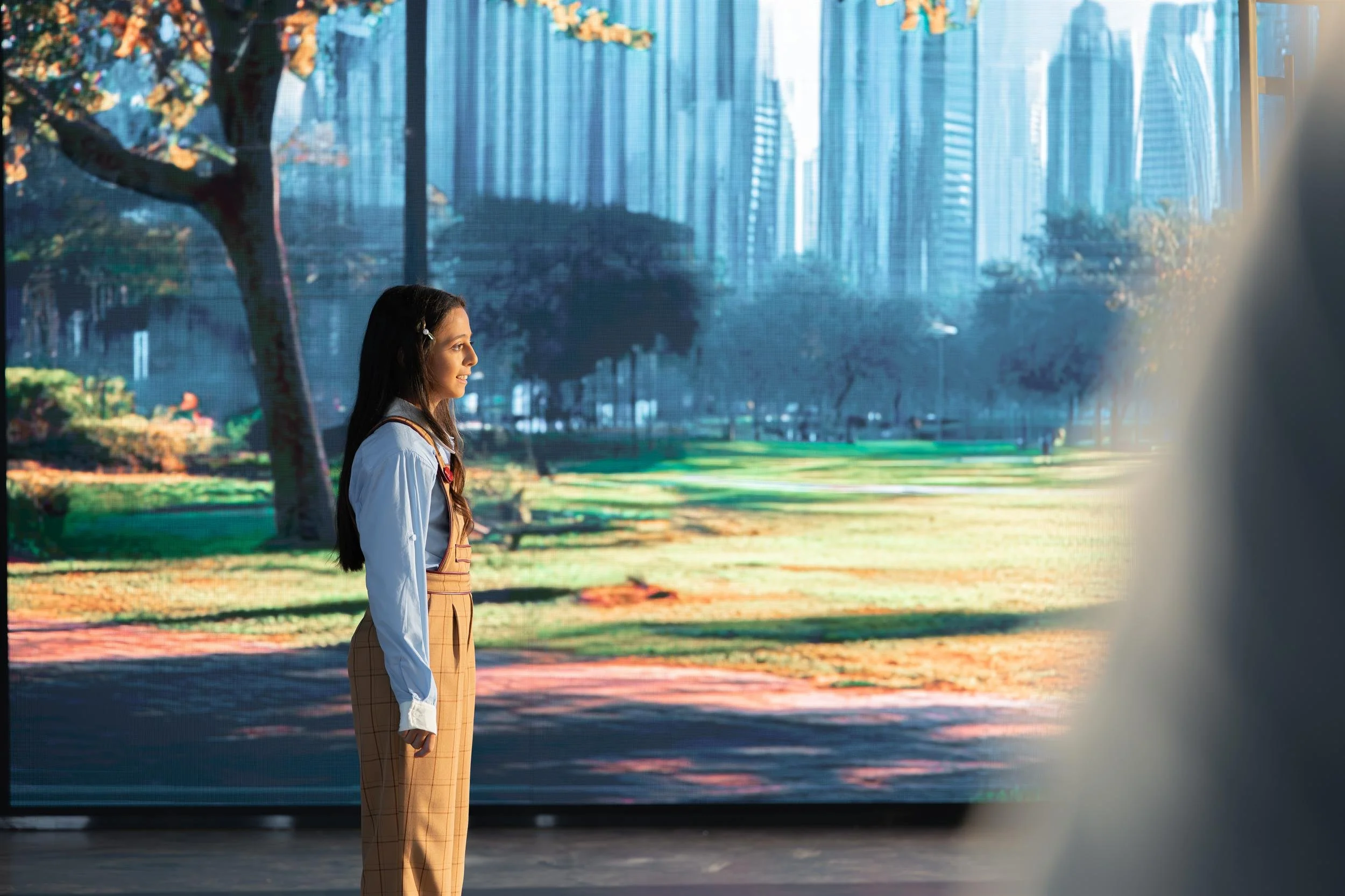 Ahlan Simsim unit still: A young girl stands in a studio in front of a large screen showing a park and city skyline.