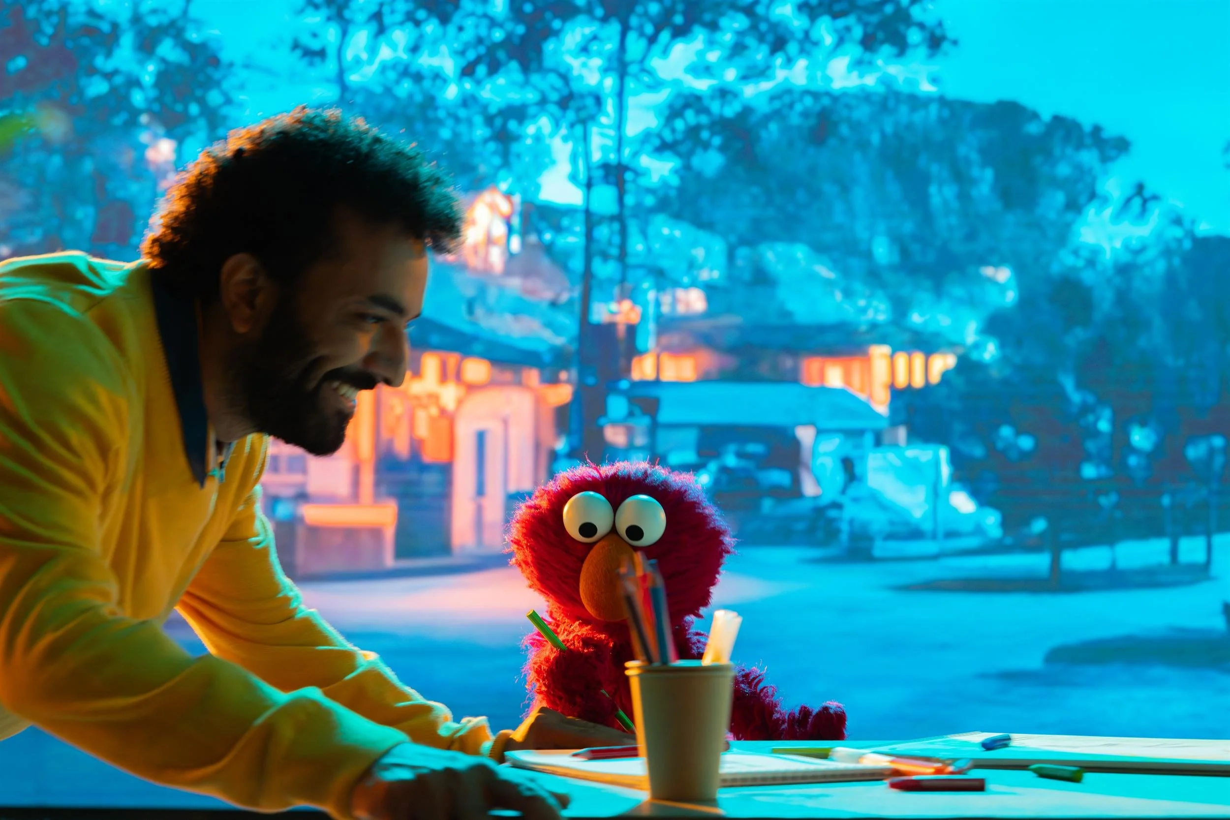 Ahlan Simsim unit still: a man in a yellow sweater leans over a desk smiling at Elmo.