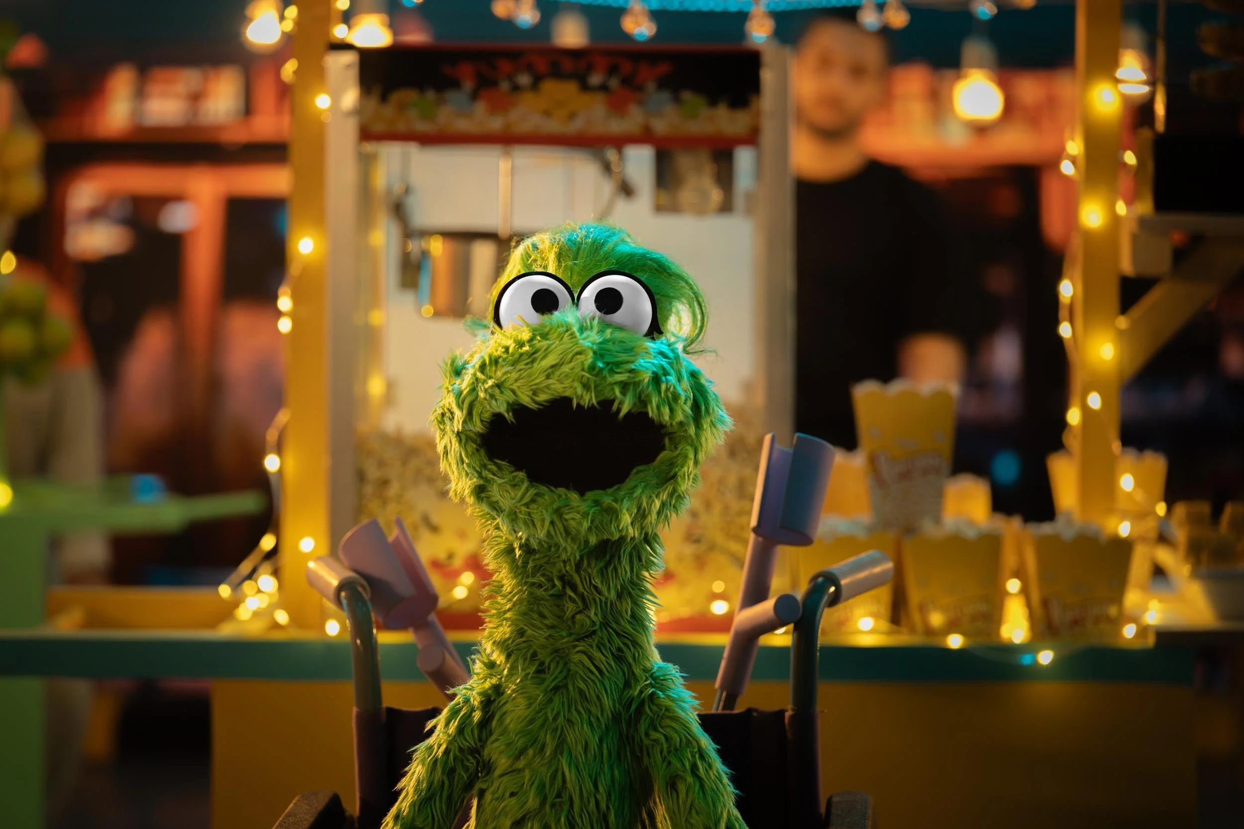 Ahlan Simsim key art: A shaggy green Muppet sits in a wheelchair in front of a brightly lit popcorn stand at night.