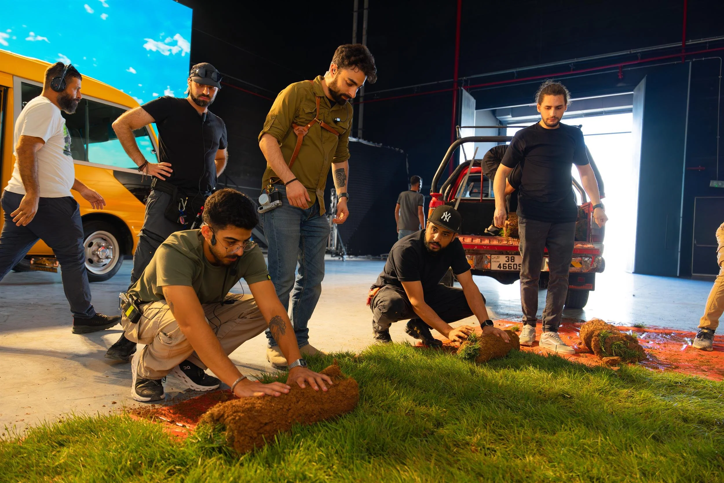 Ahlan Simsim unit still: crew members roll green sod onto a studio floor beside a yellow bus and red truck.