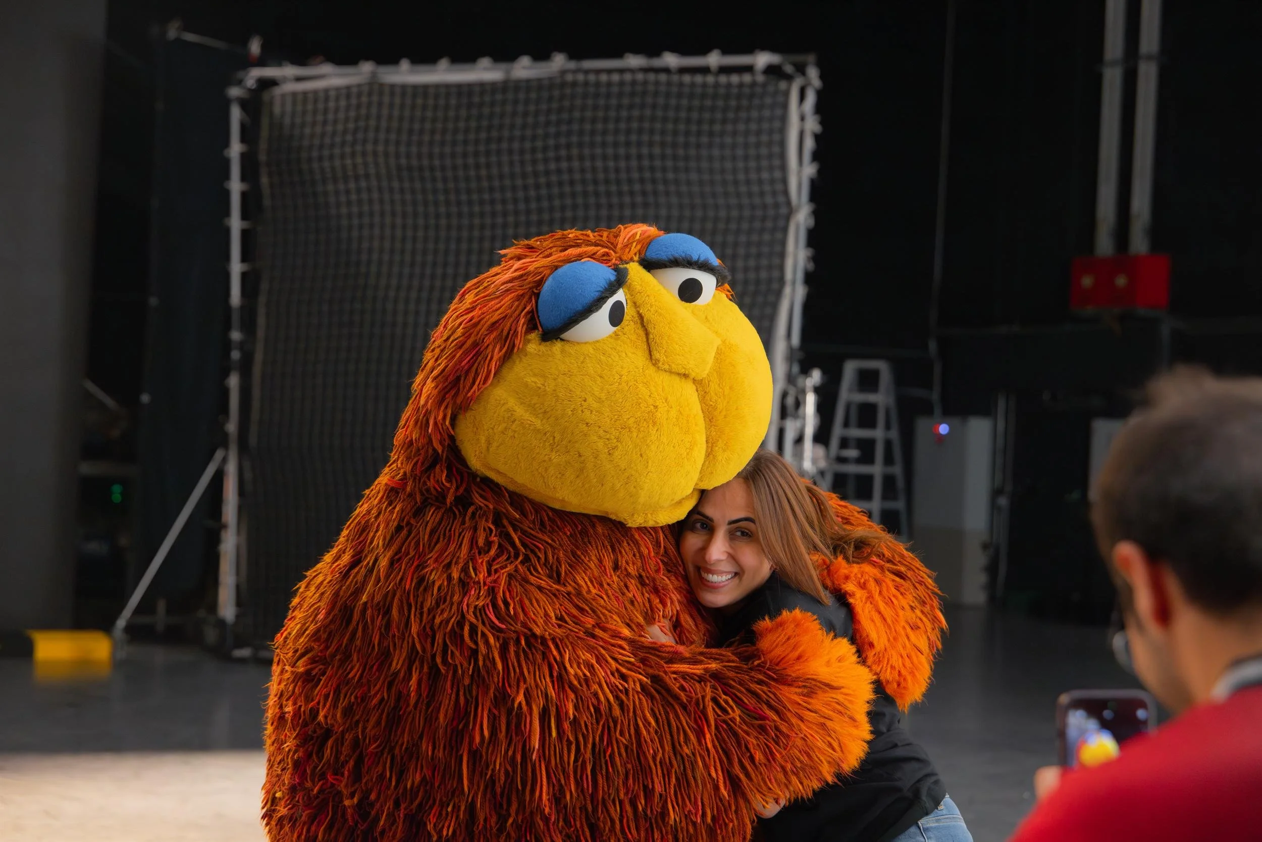 Ahlan Simsim unit still: a woman smiles while hugging a large shaggy brown Muppet on a studio set.