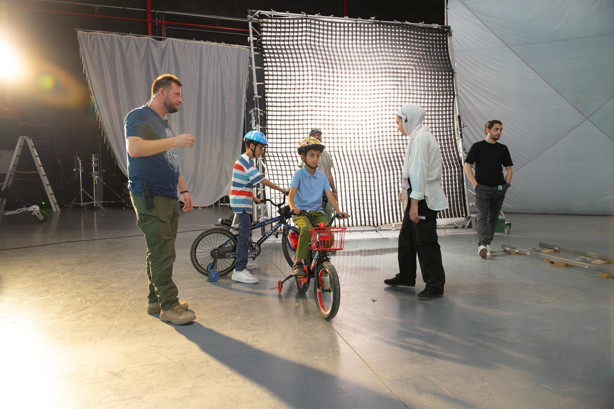Ahlan Simsim unit still: crew instruct two children on bicycles in front of a large lighting diffuser.