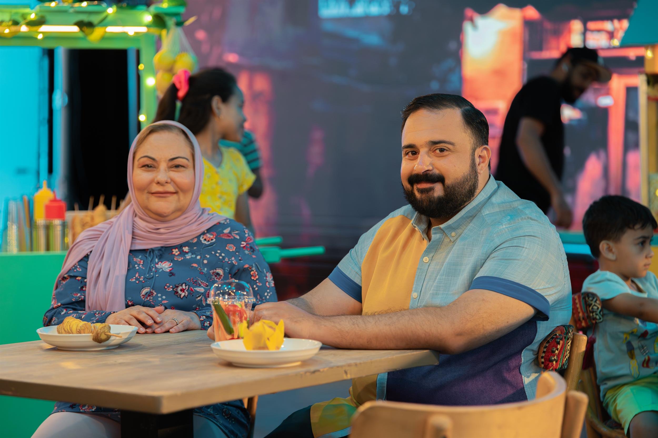 Ahlan Simsim unit still: a woman and a man sit at a table with snacks on a colorful night-market set.