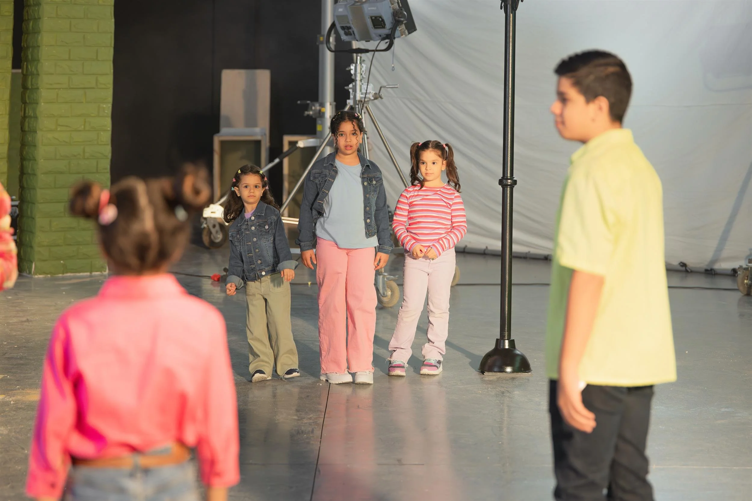 Ahlan Simsim unit still: Three girls stand on a studio floor with production lights and a white backdrop behind them.