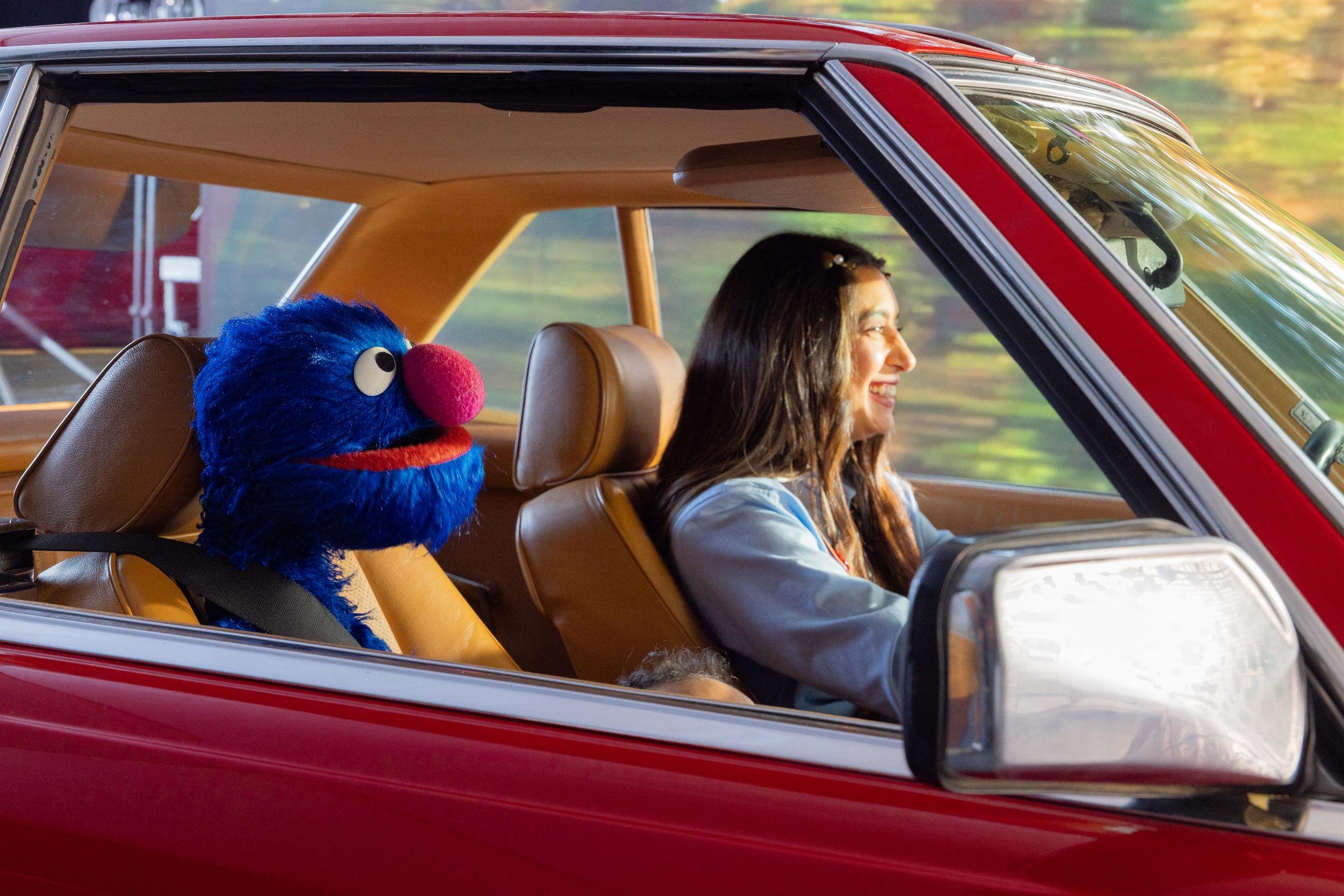 Ahlan Simsim unit still: a blue Muppet sits belted in the back of a vintage red car while a woman smiles in front.