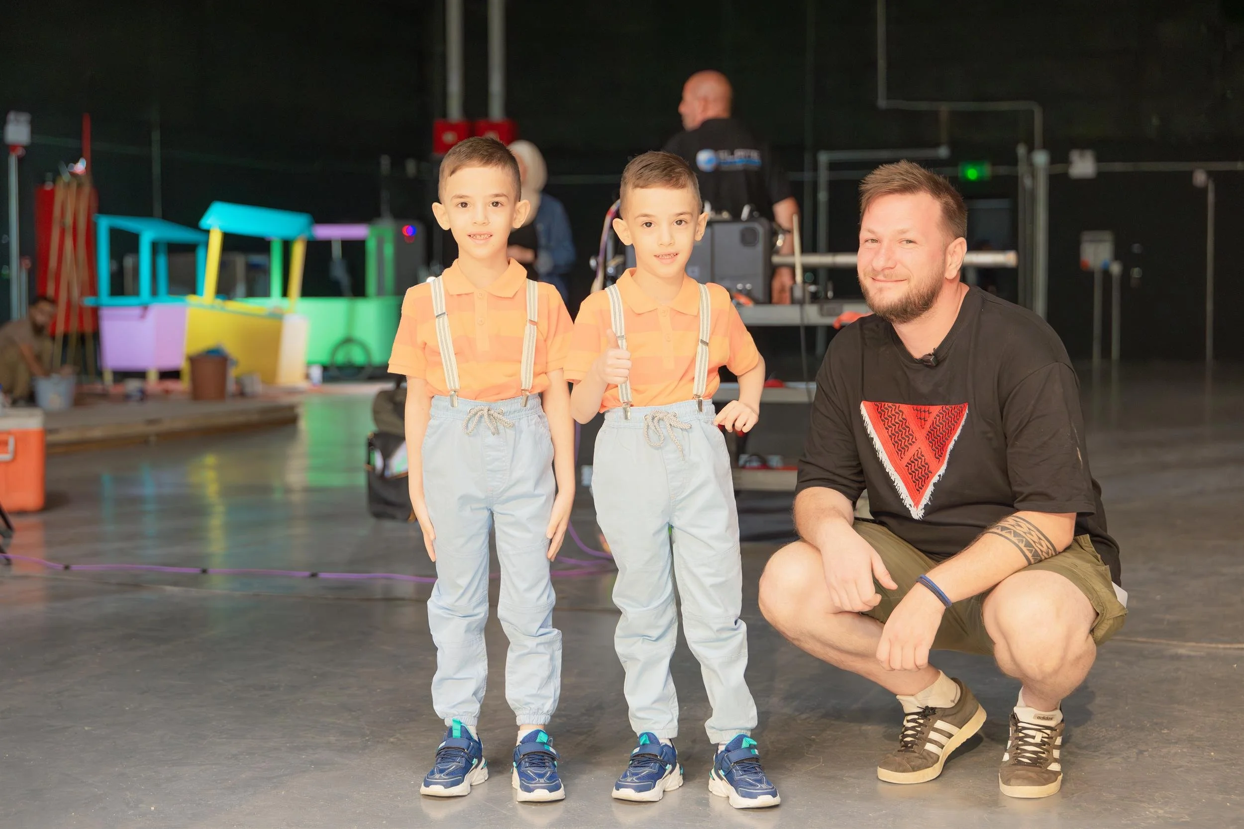 Ahlan Simsim unit still: Two boys in matching striped shirts and suspenders stand beside a smiling crew member on set.