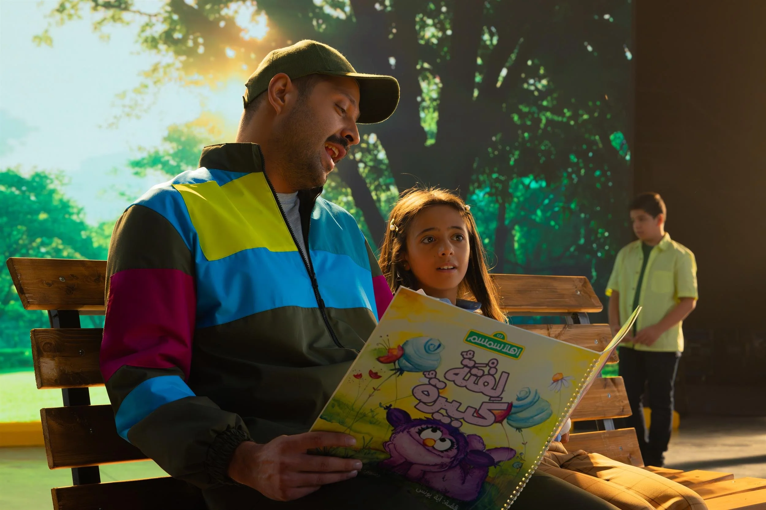 Ahlan Simsim unit still: A man in a colorful jacket reads a picture book to a young girl on a wooden park bench.