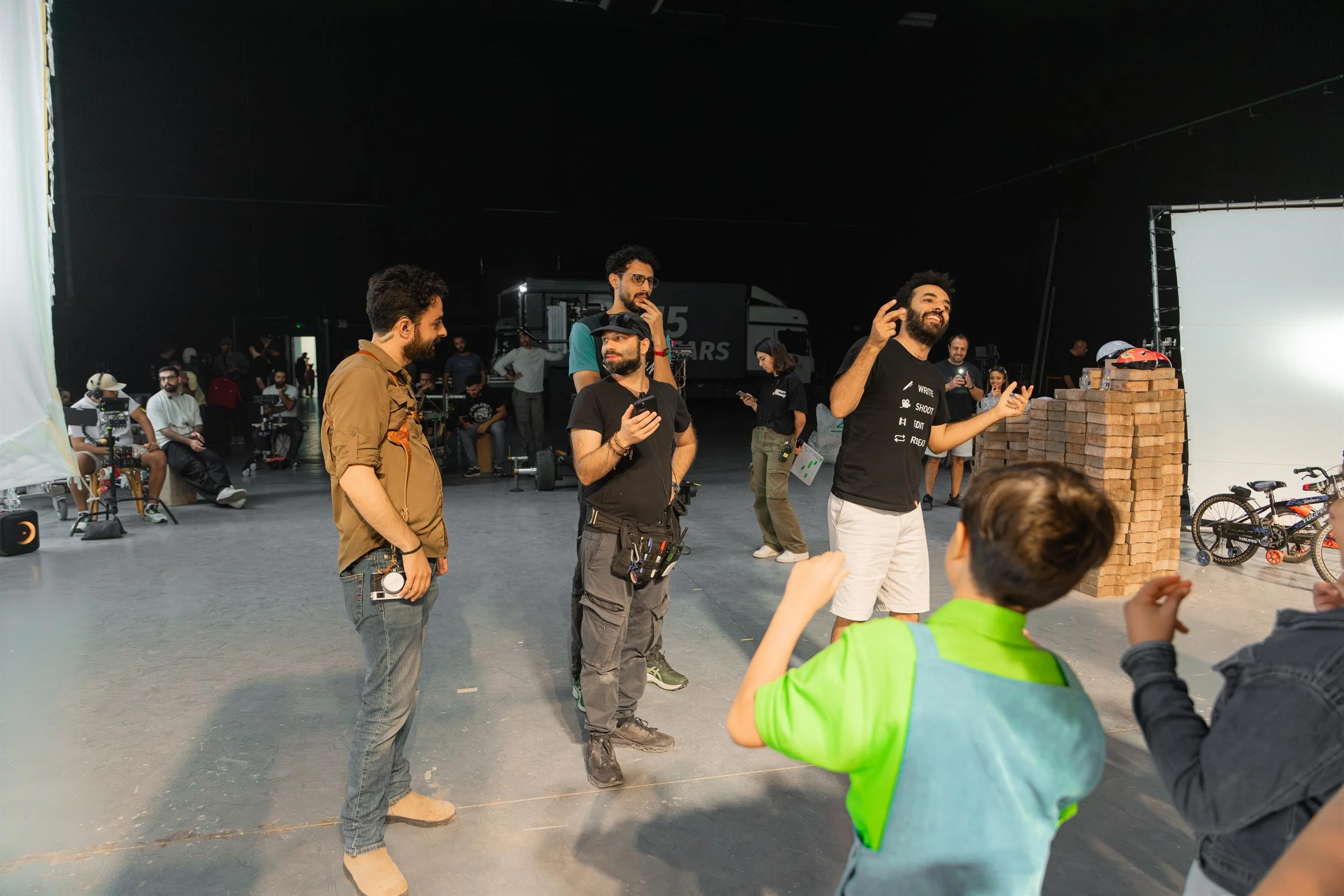 Ahlan Simsim unit still: A crew member gestures while talking with a child actor near a prop brick wall on set.