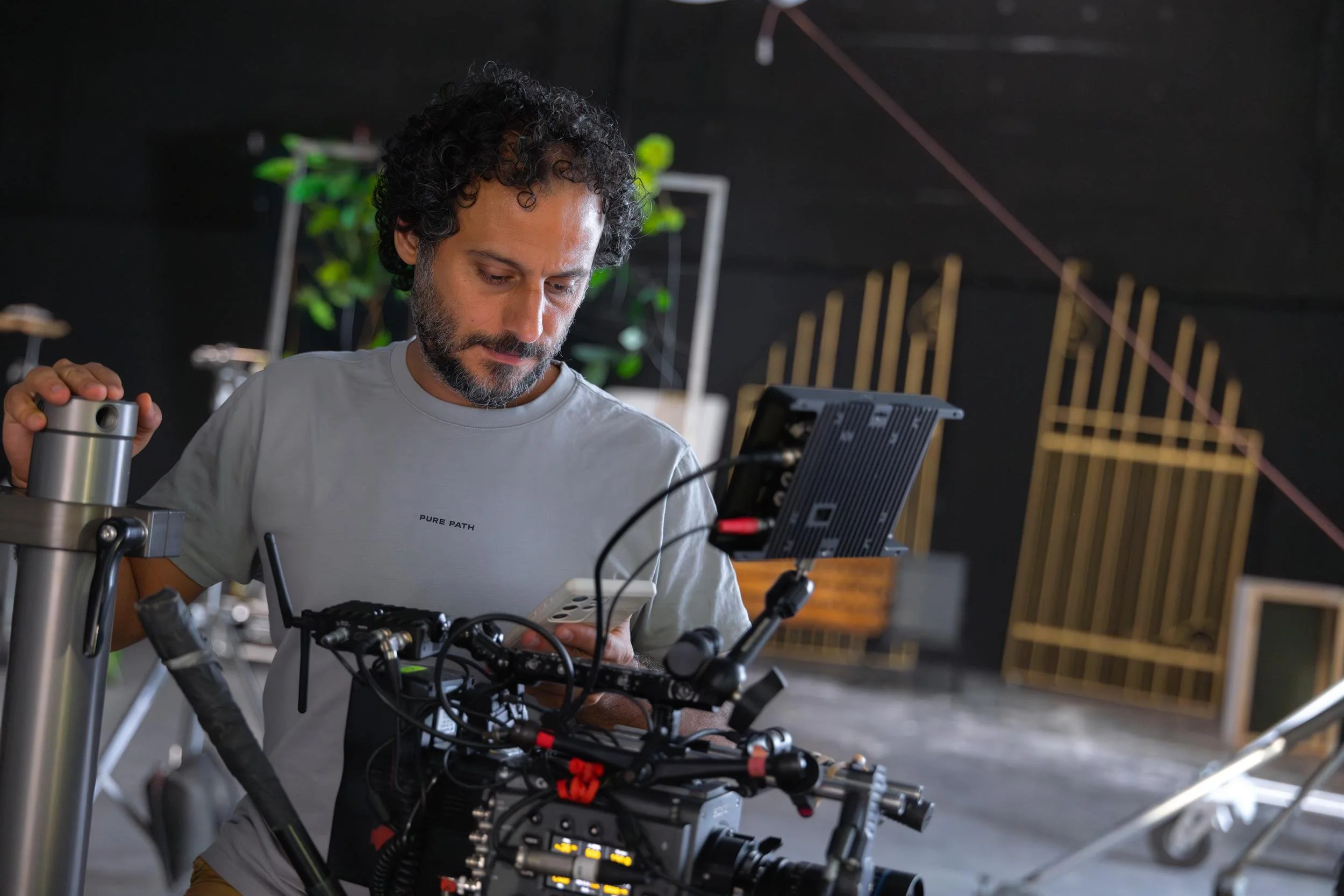 Ahlan Simsim unit still: a crew member looks at a smartphone beside a camera rig and golden park gates.