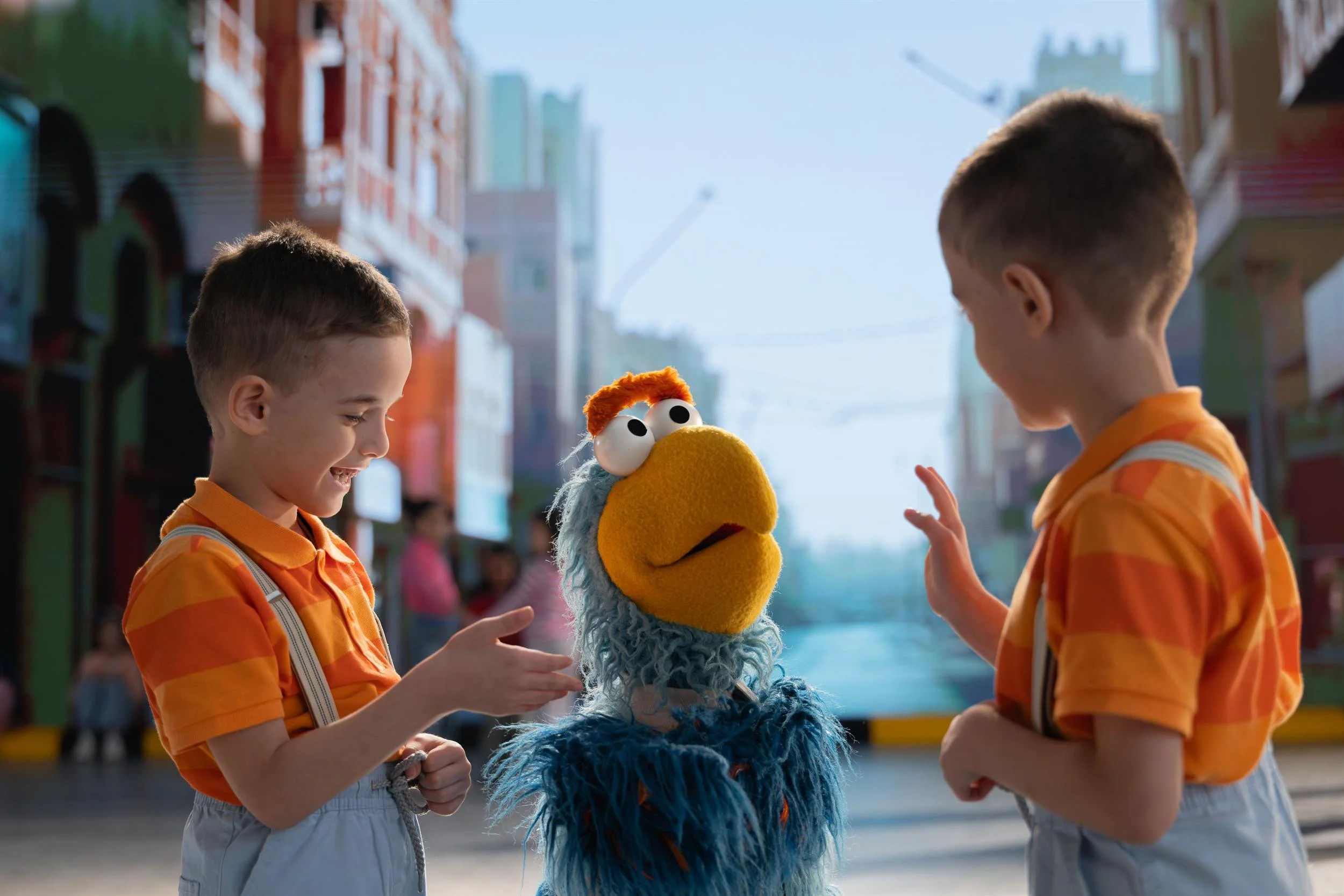 Ahlan Simsim key art: Two boys in striped shirts talk to a blue Muppet with a yellow beak on a colorful street set.