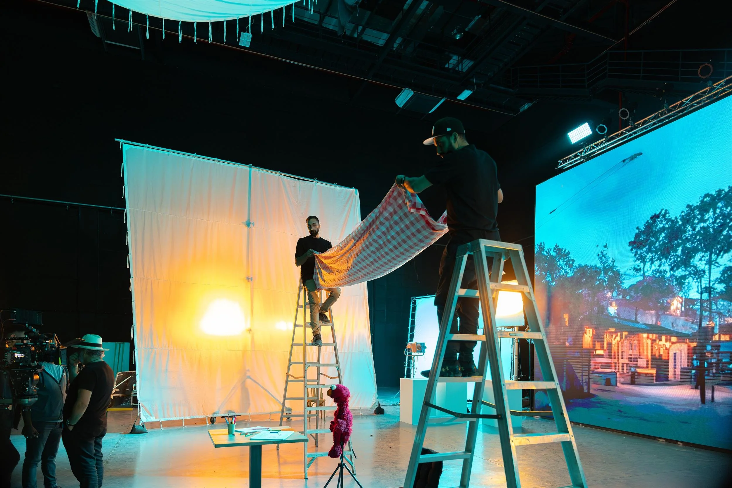Ahlan Simsim unit still: crew on ladders hold a checkered cloth while Elmo sits near a table and an LED street screen.