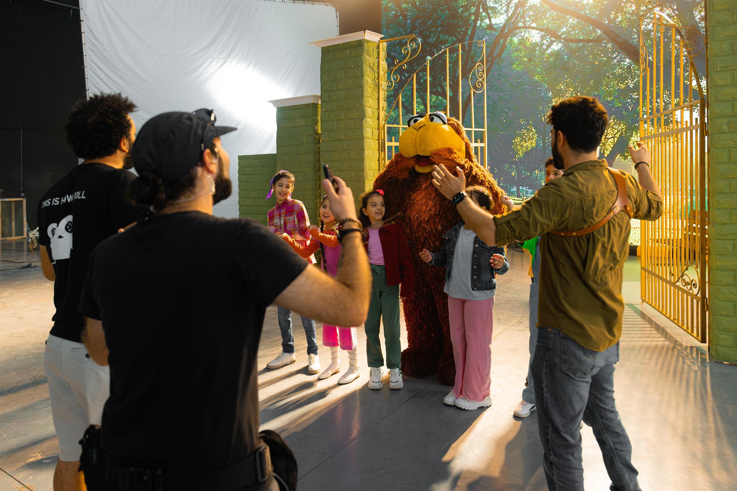 Ahlan Simsim unit still: Crew film a large brown Muppet and three children in front of golden gates and a park backdrop.