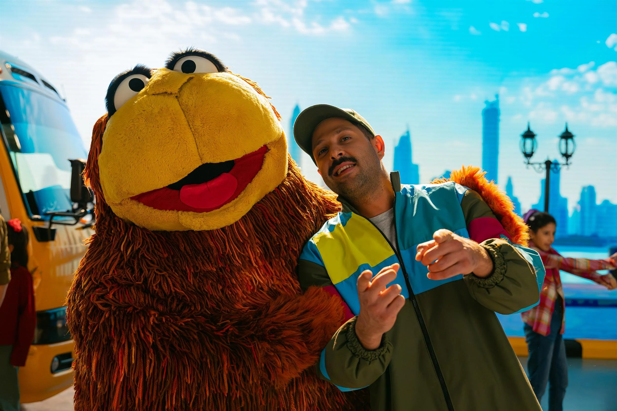 Ahlan Simsim unit still: a man in a colorful jacket poses with a large brown Muppet in front of a city skyline.