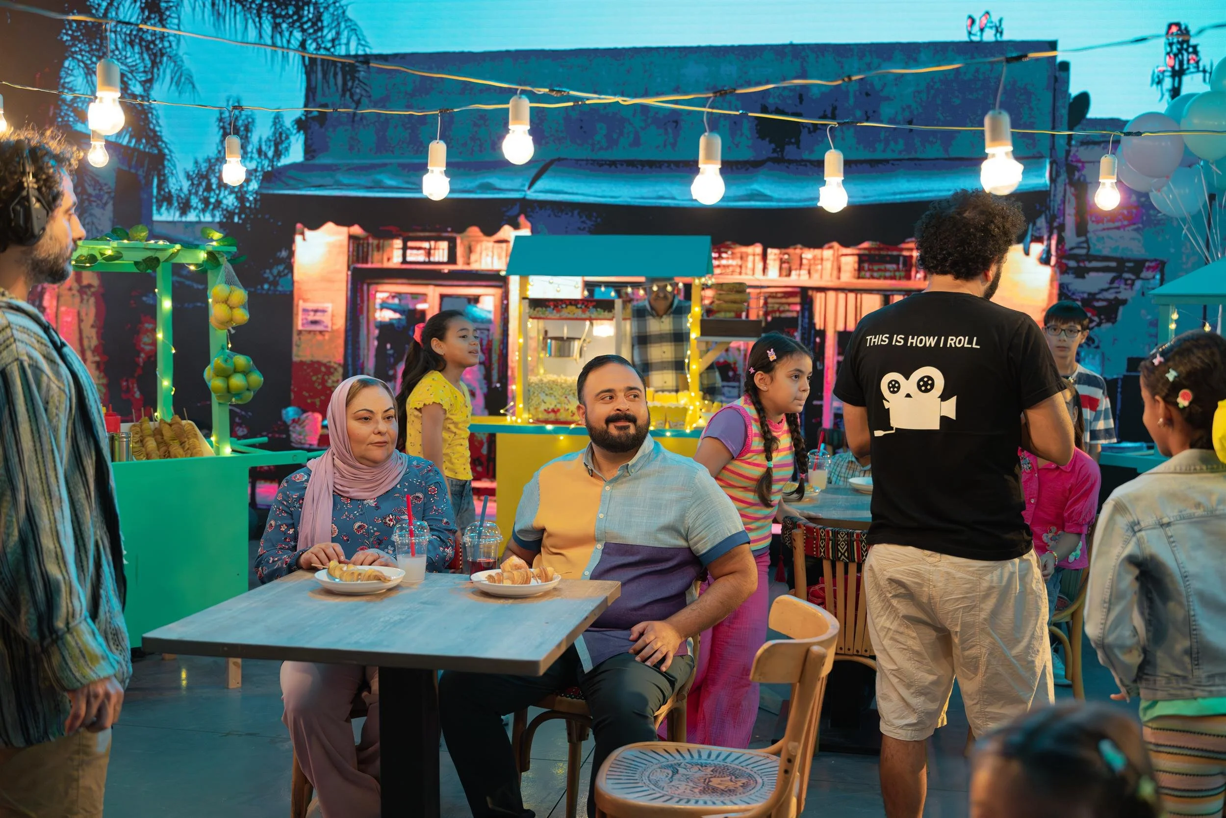 Ahlan Simsim unit still: crew and actors on a colorful night-market set with string lights and food stalls.