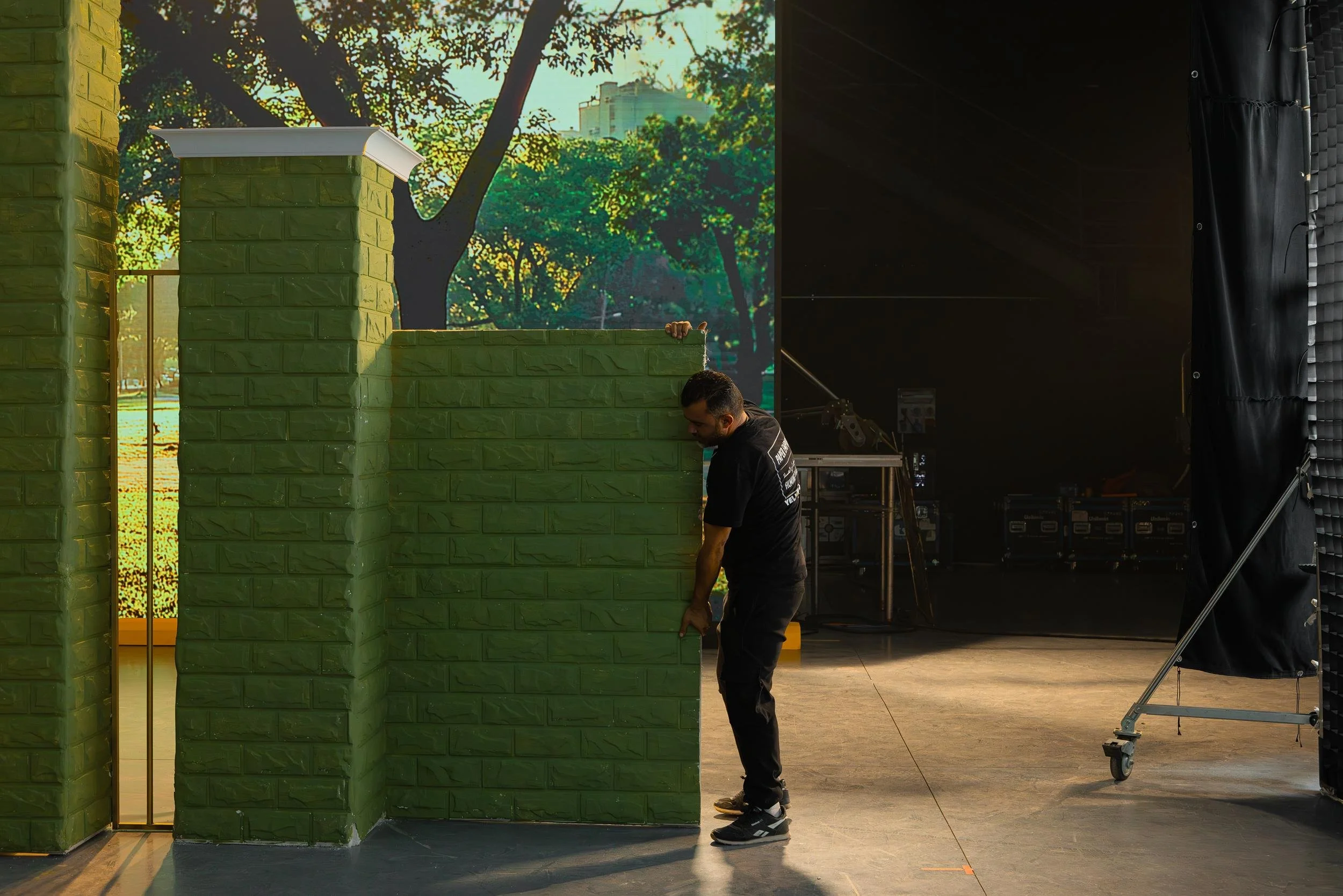 Ahlan Simsim unit still: A crew member moves a large green brick-textured set piece in front of a park backdrop.