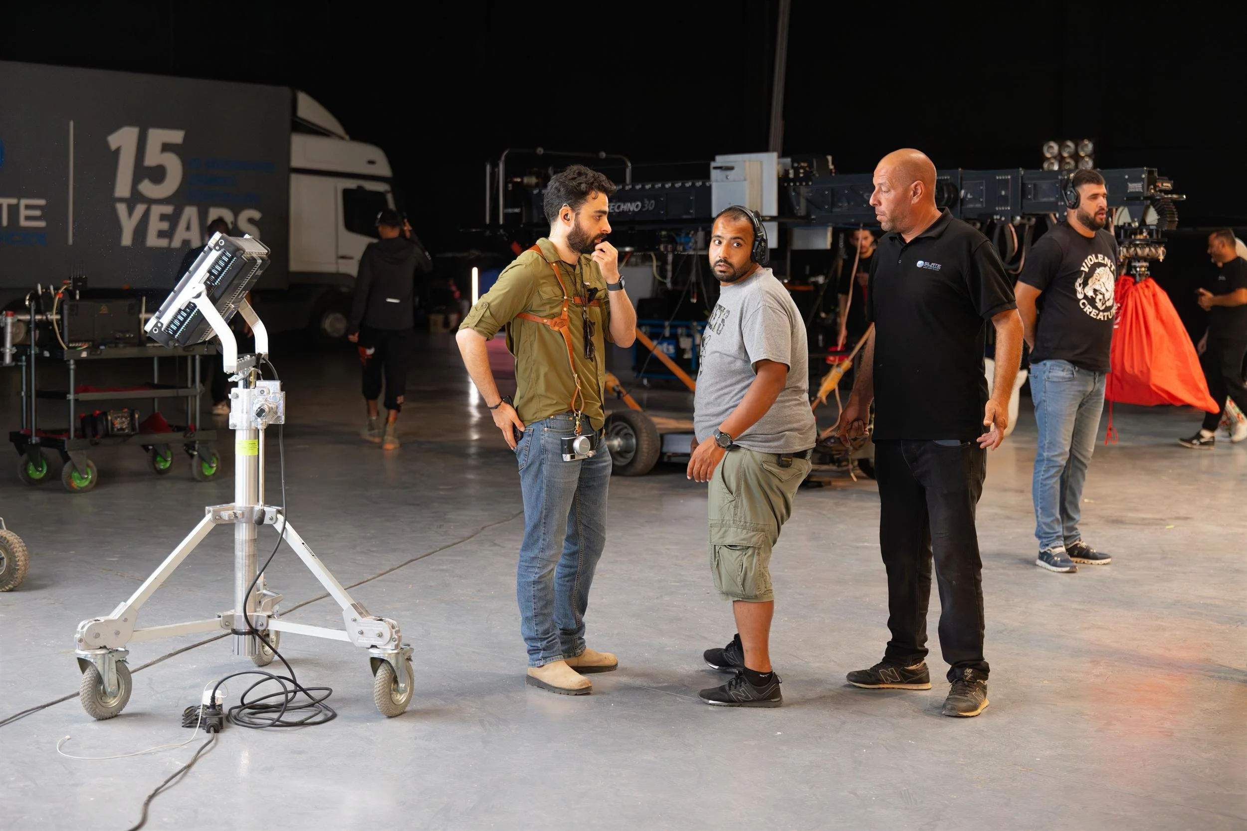 Ahlan Simsim unit still: Three crew members talk in a large studio with filming equipment in the background.