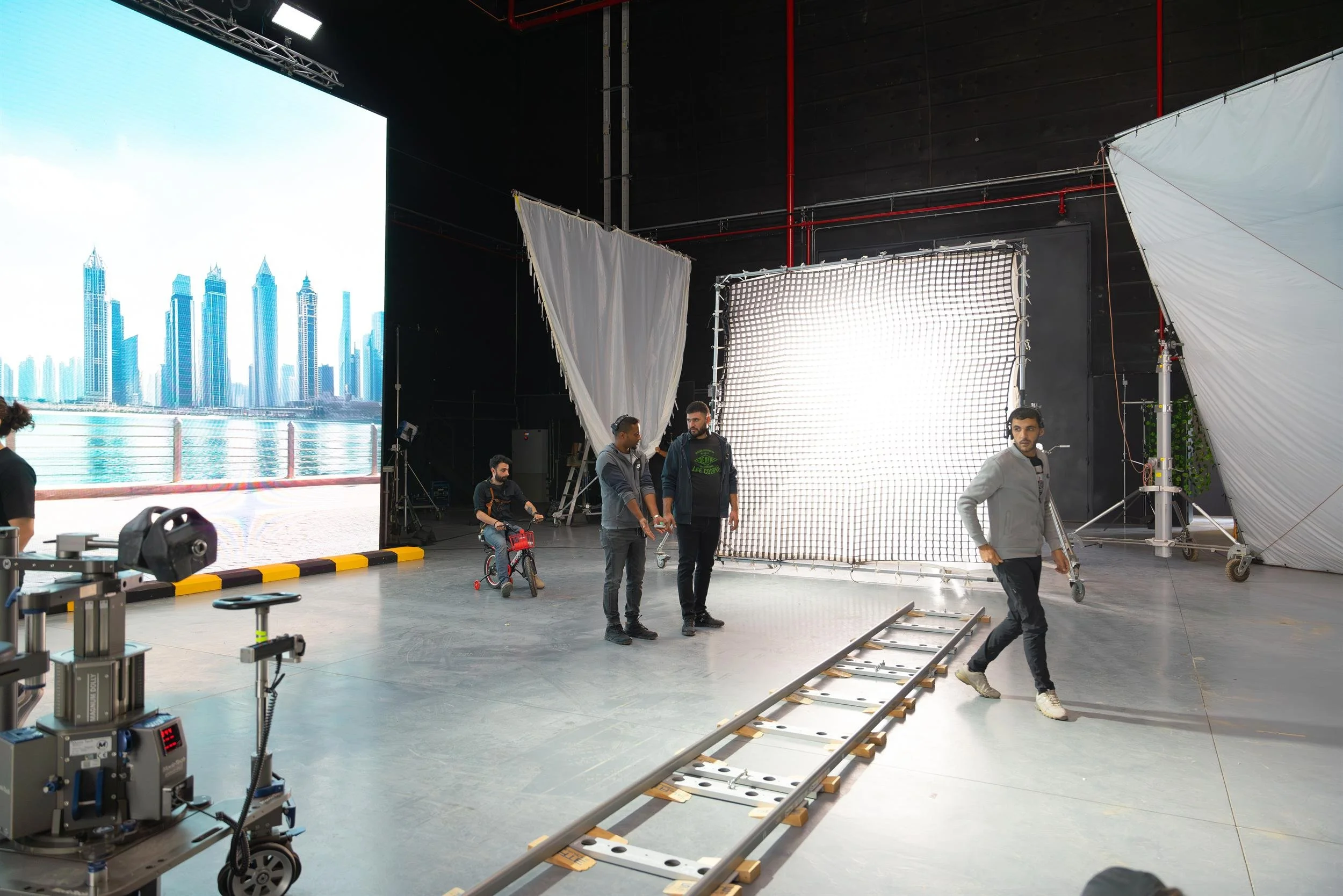 Ahlan Simsim unit still: crew talk near camera tracks and a red bicycle in front of an LED city skyline.
