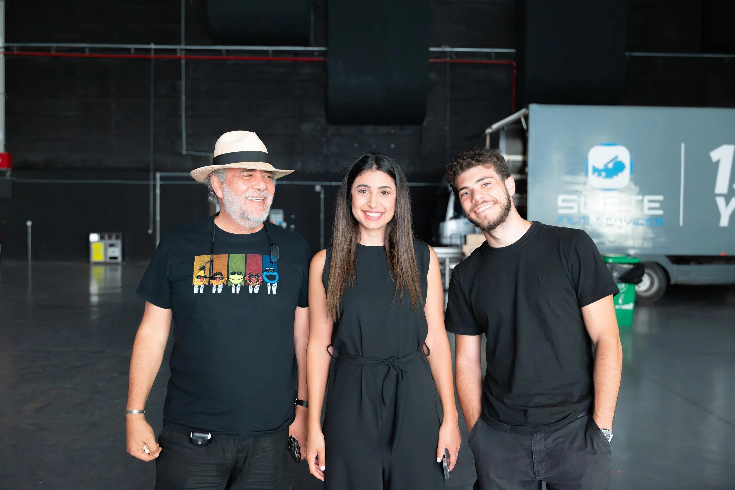Ahlan Simsim unit still: three smiling crew members pose in a dark studio with a production truck behind them.