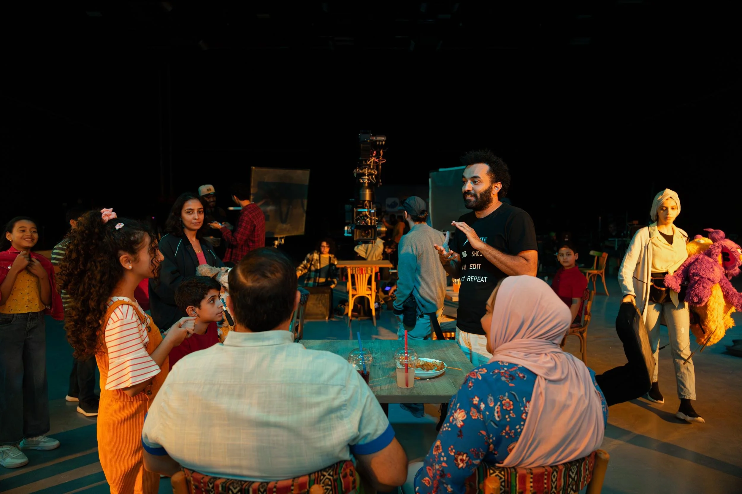 Ahlan Simsim unit still: a crew member talks to children and adults at a table while a woman holds a purple Muppet.