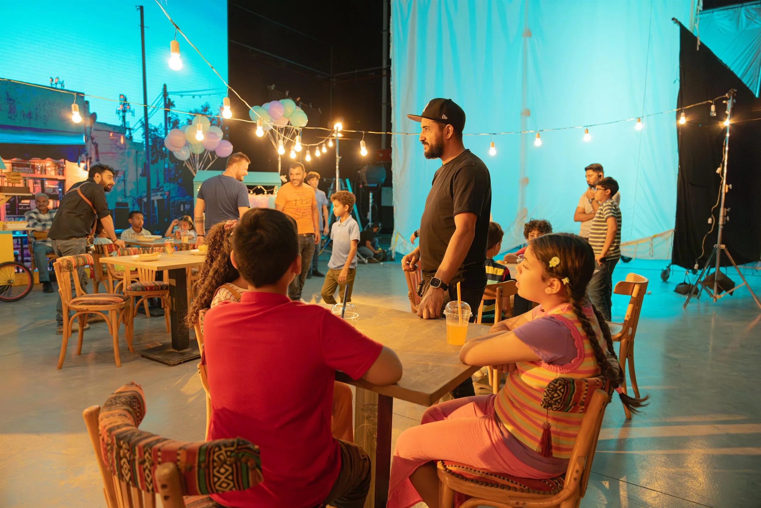 Ahlan Simsim unit still: children sit at cafe tables on a night-market set with string lights and crew nearby.