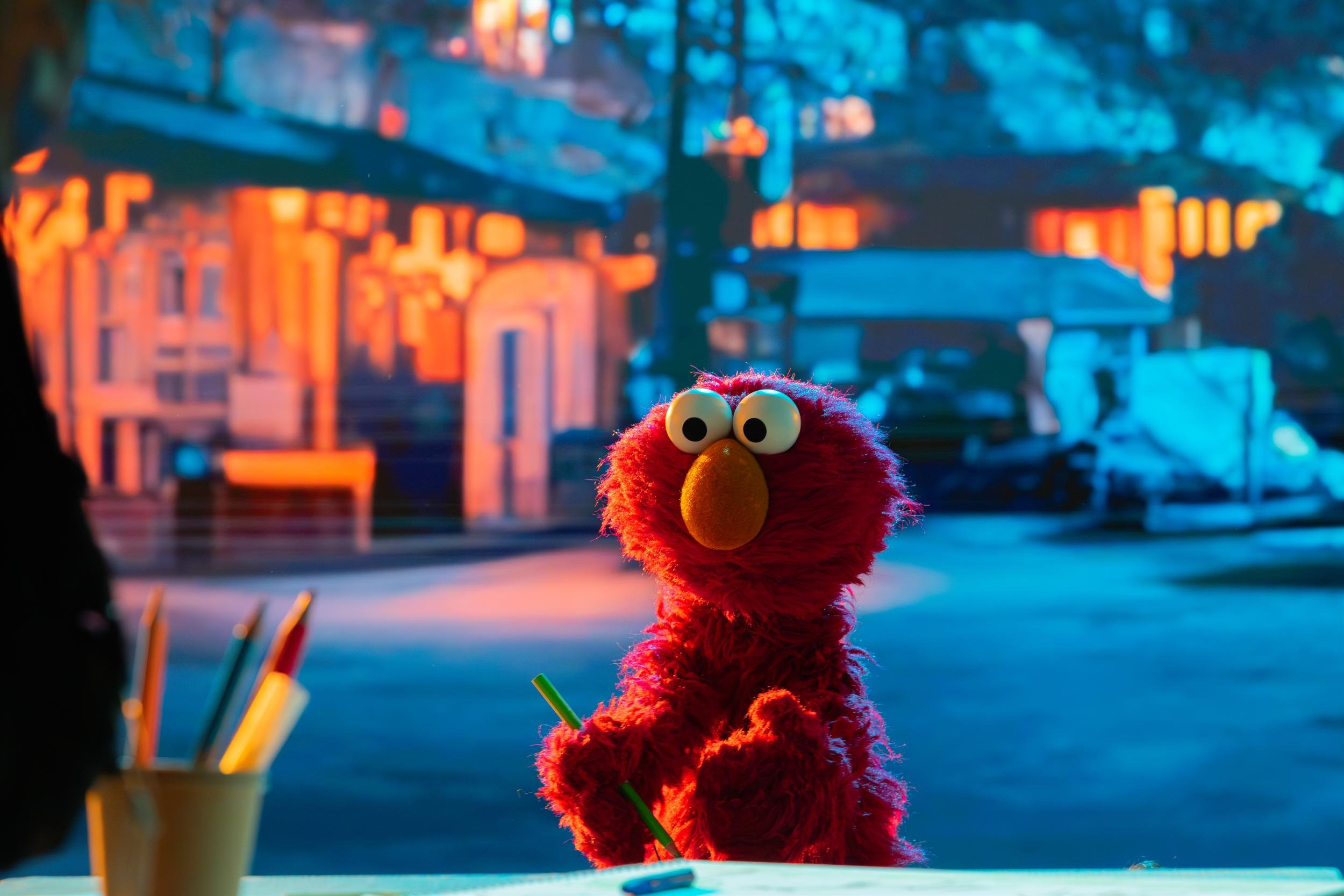 Ahlan Simsim unit still: Elmo holds a green pencil at a desk against a blurred night street.