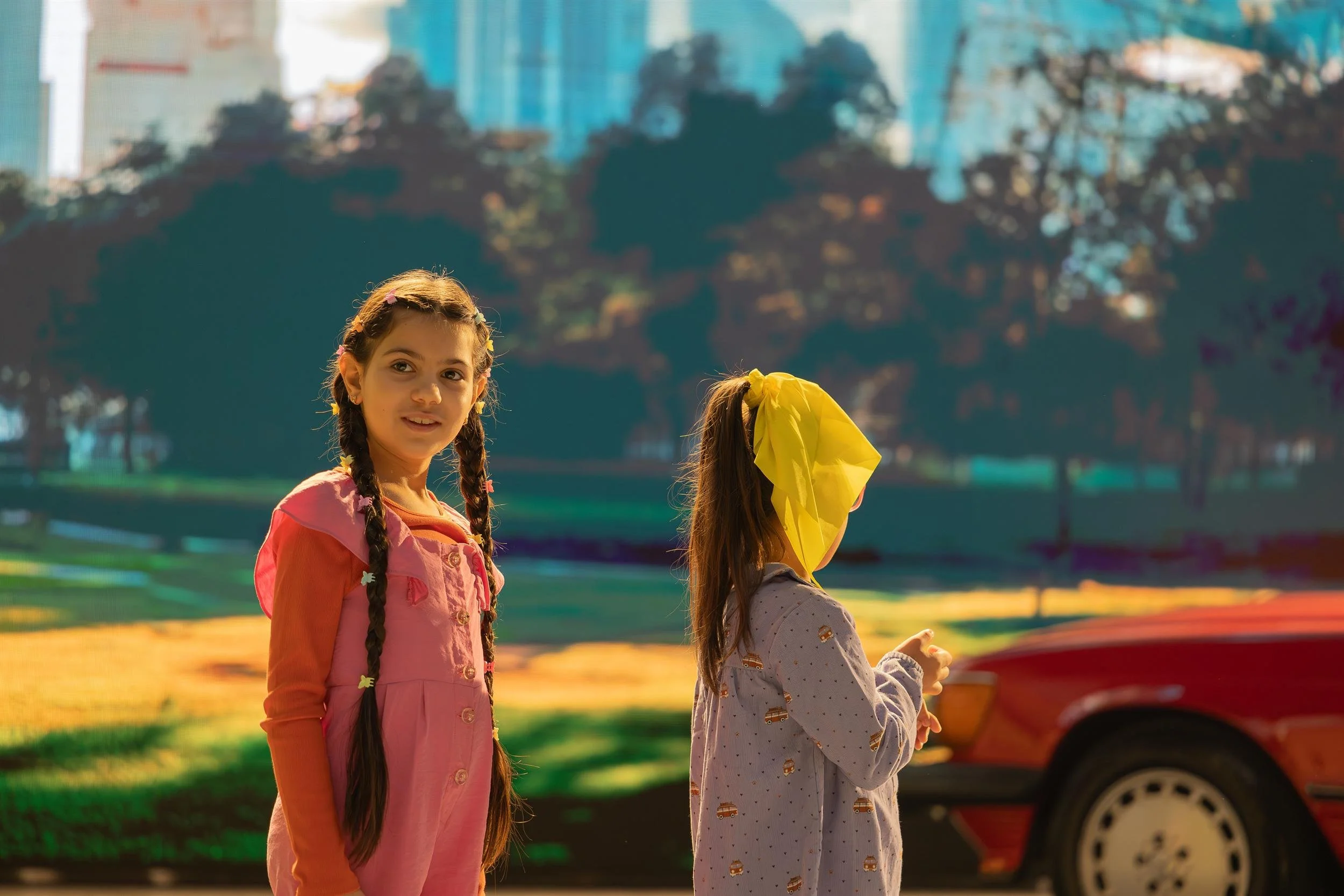 Ahlan Simsim unit still: Two young girls stand in front of a park backdrop with a red car nearby.