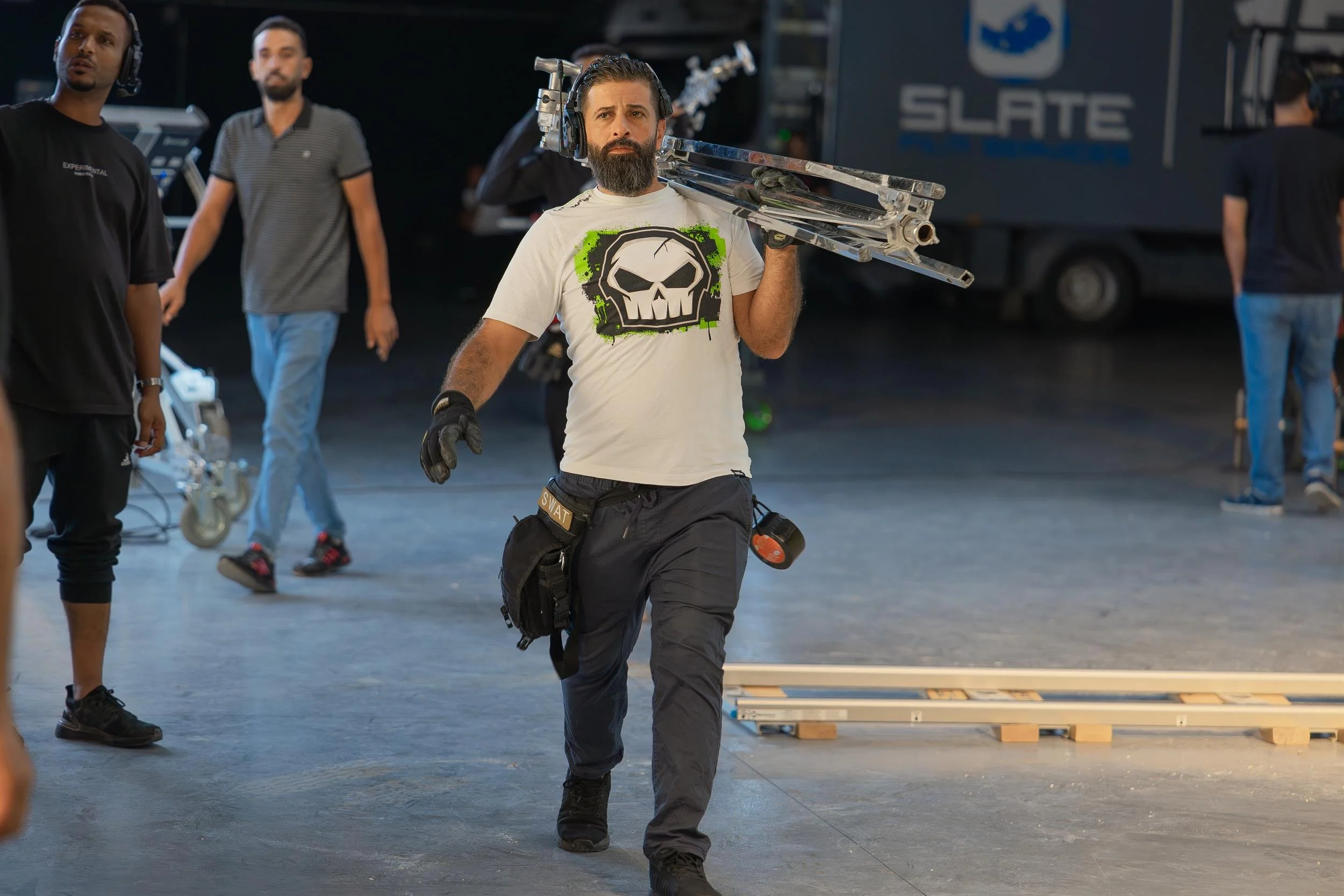 Ahlan Simsim unit still: A bearded crew member carries a large tripod across a studio floor during production.