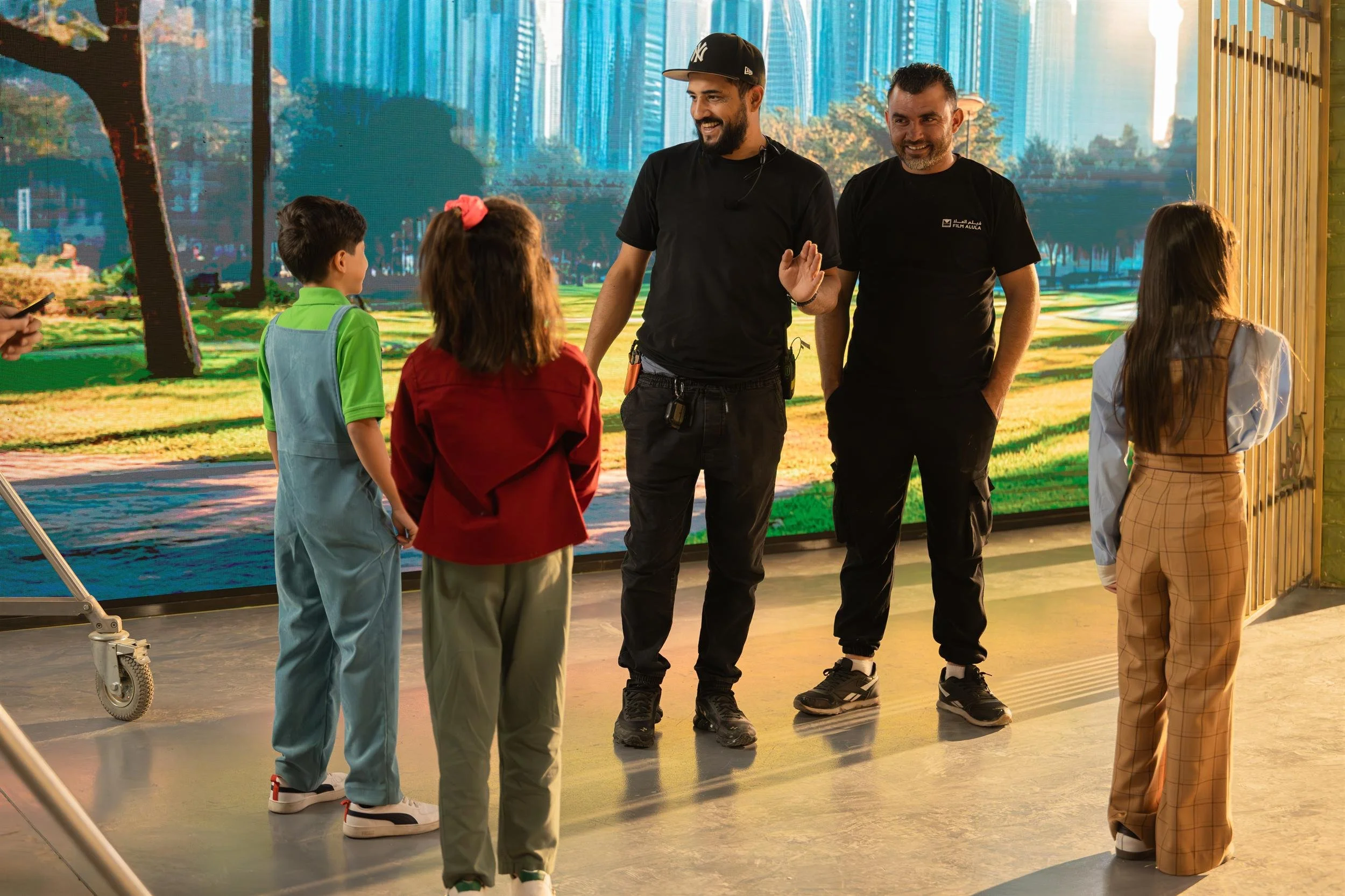 Ahlan Simsim unit still: Two crew members talk with three children on a studio set in front of a park LED wall.