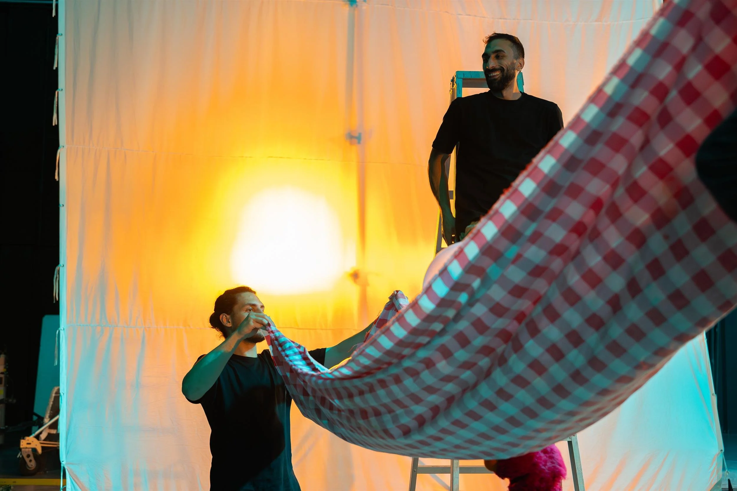 Ahlan Simsim unit still: two crew members hold a red-and-white checkered cloth beside a ladder and orange-lit scrim.