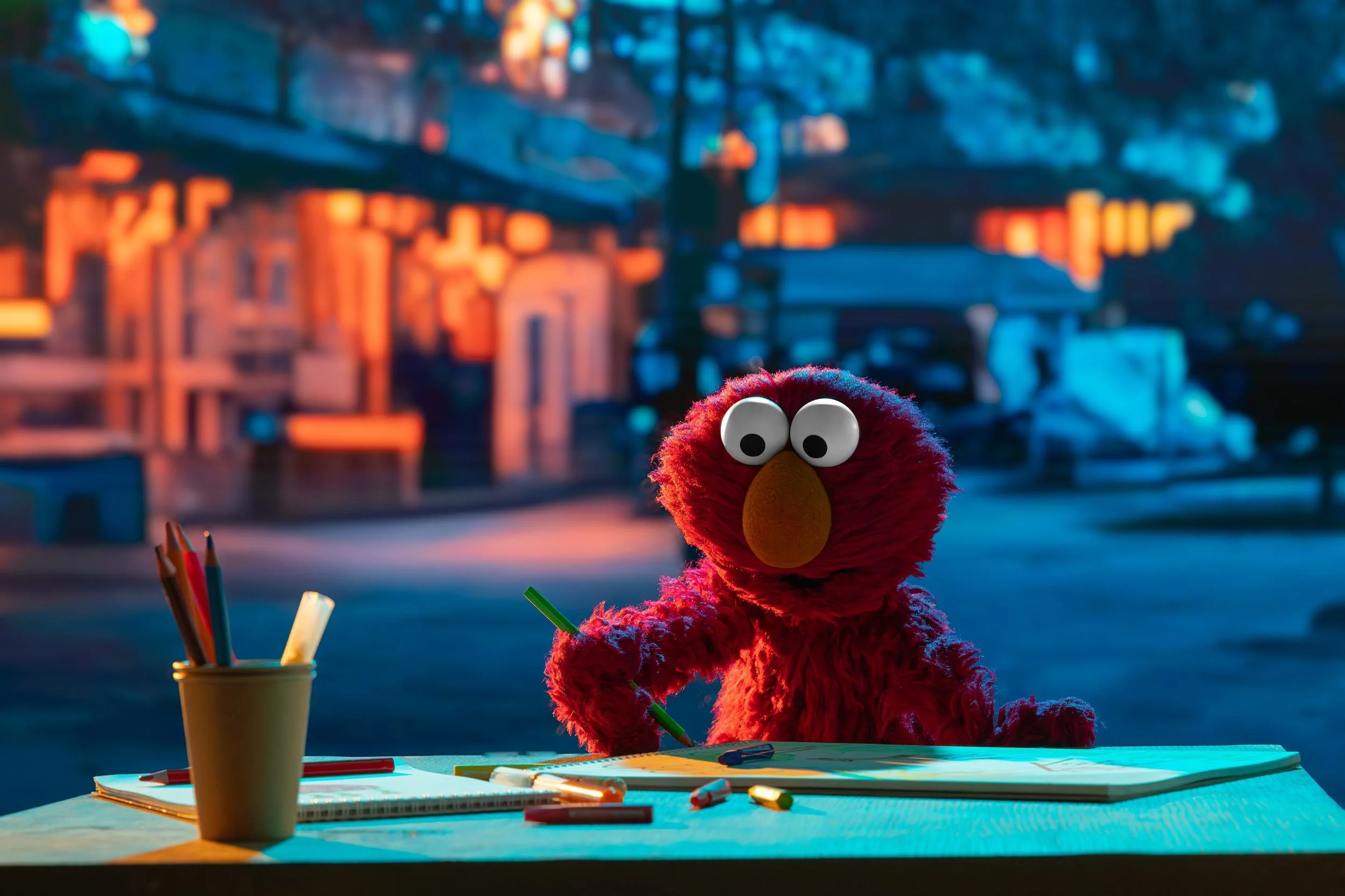 Ahlan Simsim key art: Elmo draws at an outdoor table at night with pencils and a sketchbook.