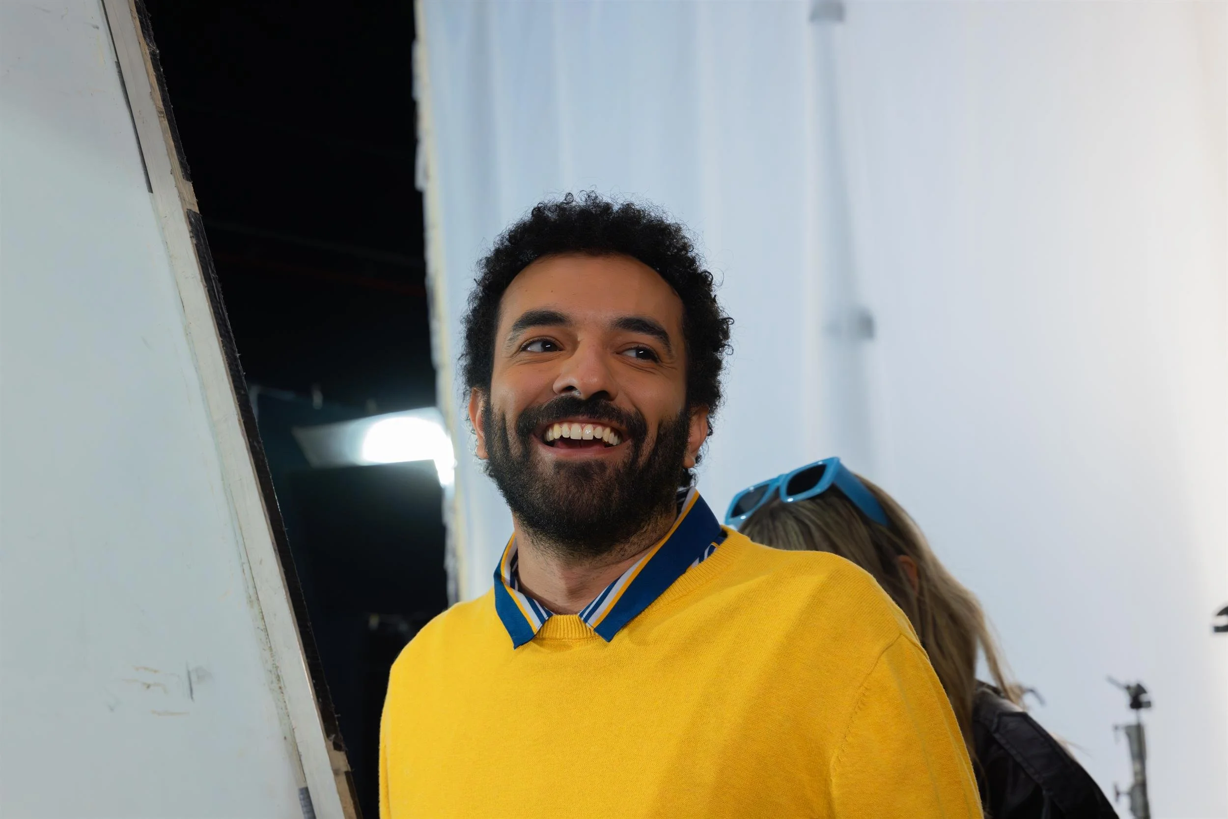 Ahlan Simsim unit still: a smiling man in a yellow sweater looks off-camera on a studio set.