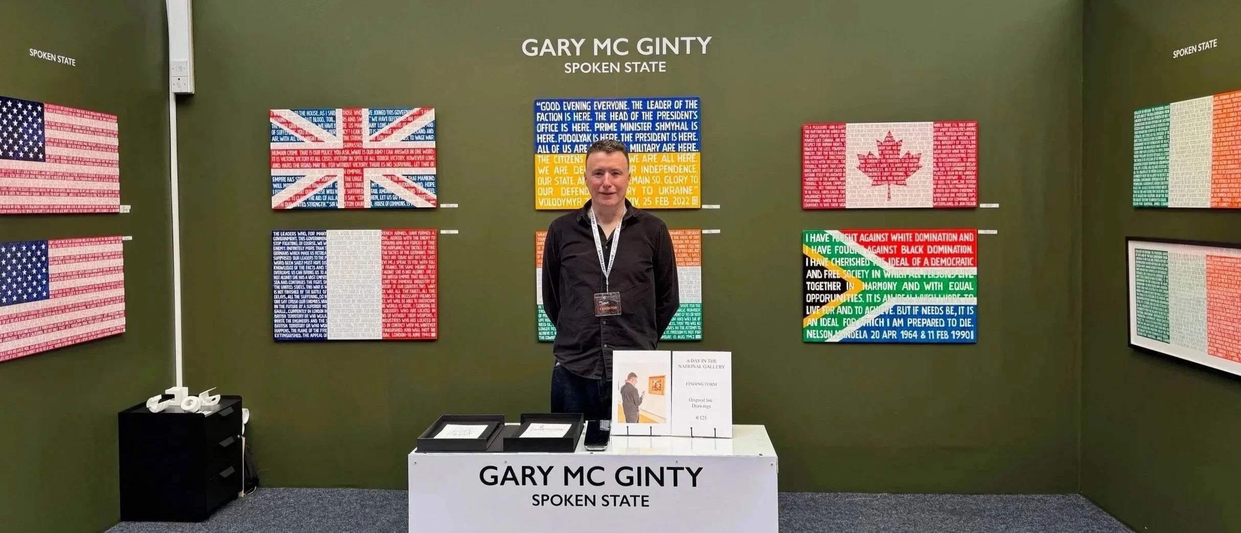 A man standing inside an art exhibition or gallery. Behind him are colorful art pieces based on the American flag, Canadian flag, and other national symbols, created with text. The display is titled 'Gary MC Ginty Spoken State'. The man is wearing a black shirt and has a lanyard around his neck. In front of him is a table with artwork cards and a sign indicating it is related to original ink drawings priced at 125.