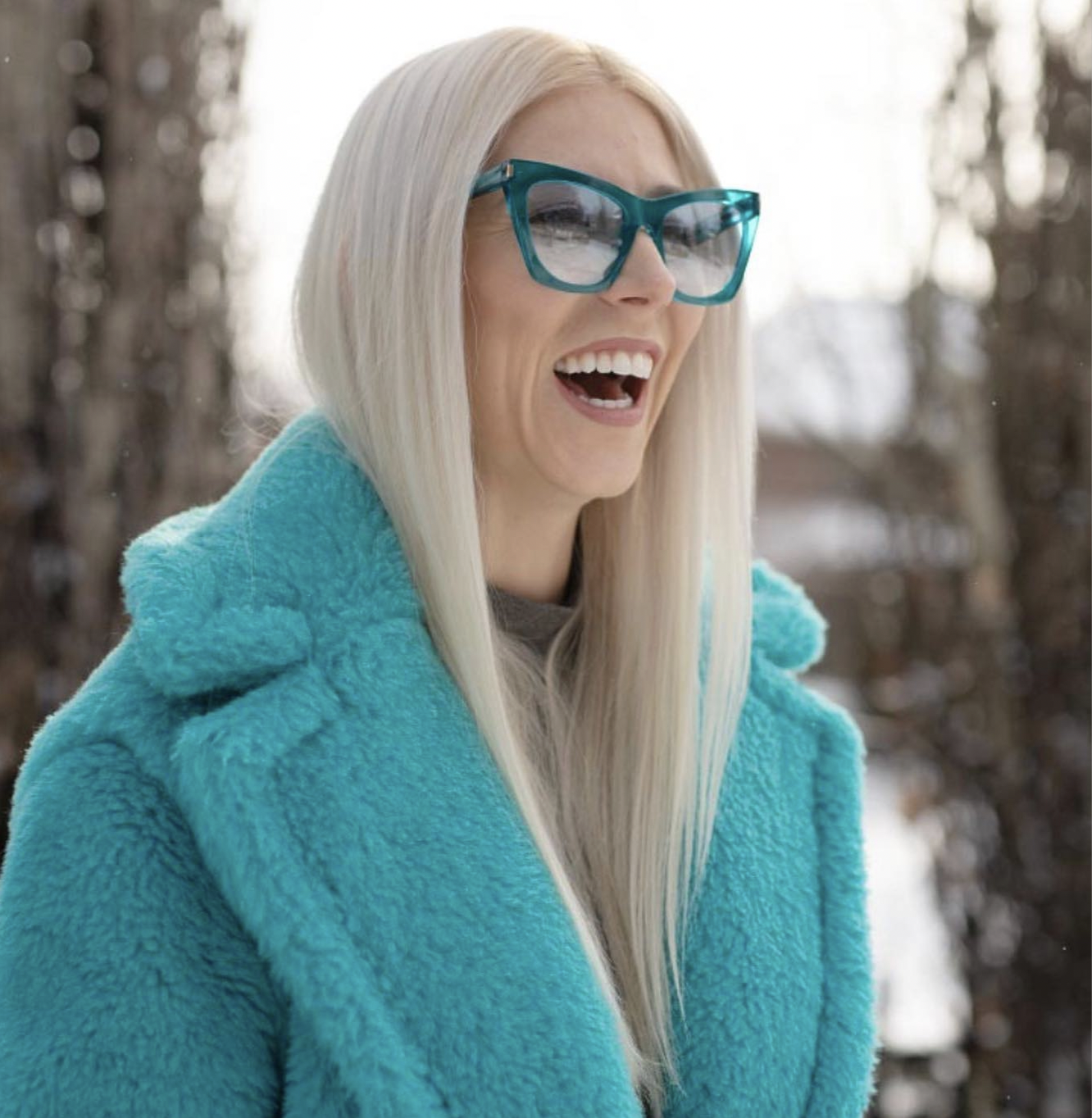 A woman with long blonde hair wearing sunglasses and a turquoise coat, smiling and looking outdoors on a snowy day.
