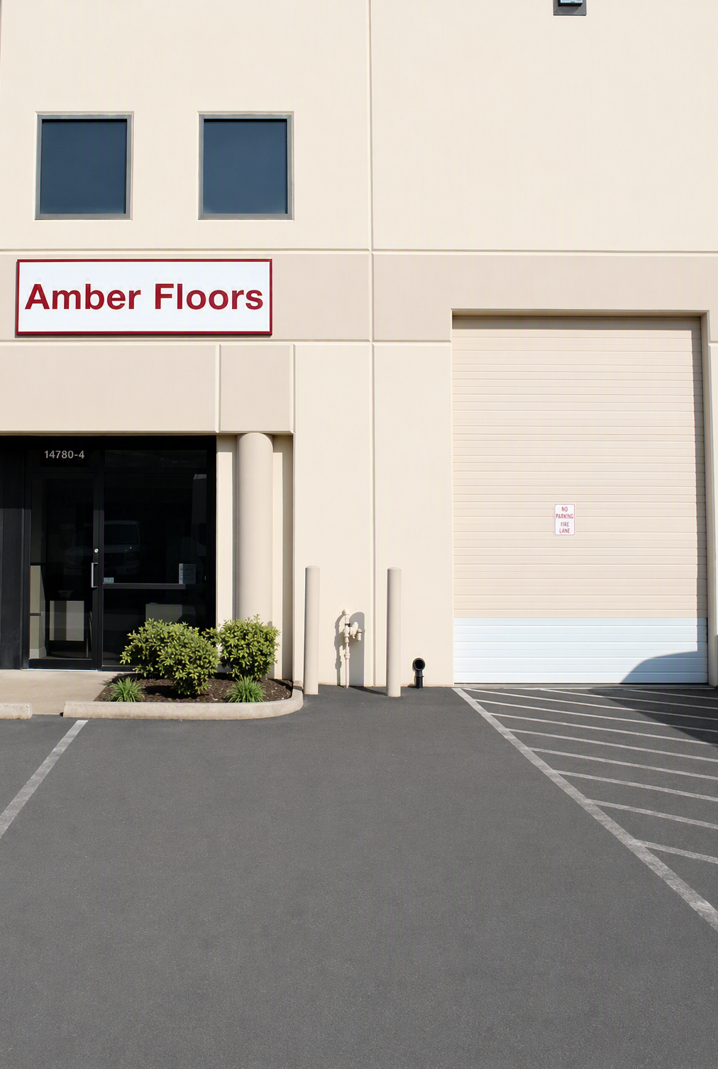 Exterior of a commercial building with a sign that reads 'Amber Floors', a glass door, a small landscaped area with bushes, a large storage door with a small sign that says 'No Parking Fire Lane', parking spaces in the foreground, and a bright, clear sky.