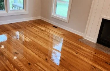 Empty room with polished, refinished hardwood floors, white walls, large windows, and a fireplace.