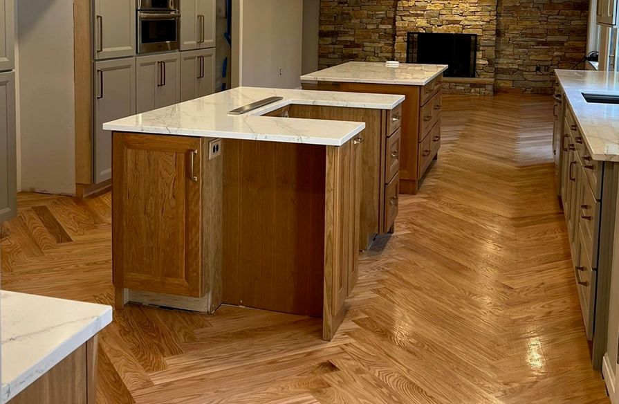 Kitchen with wooden cabinets, white marble countertops, hardwood flooring, and a stone fireplace in the background.