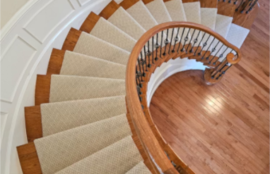 Curved staircase with beige carpeted steps and wooden handrail at the top of a wooden floor.