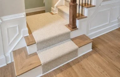 Interior staircase with beige carpeted steps, wooden handrail, and hardwood flooring at the bottom.