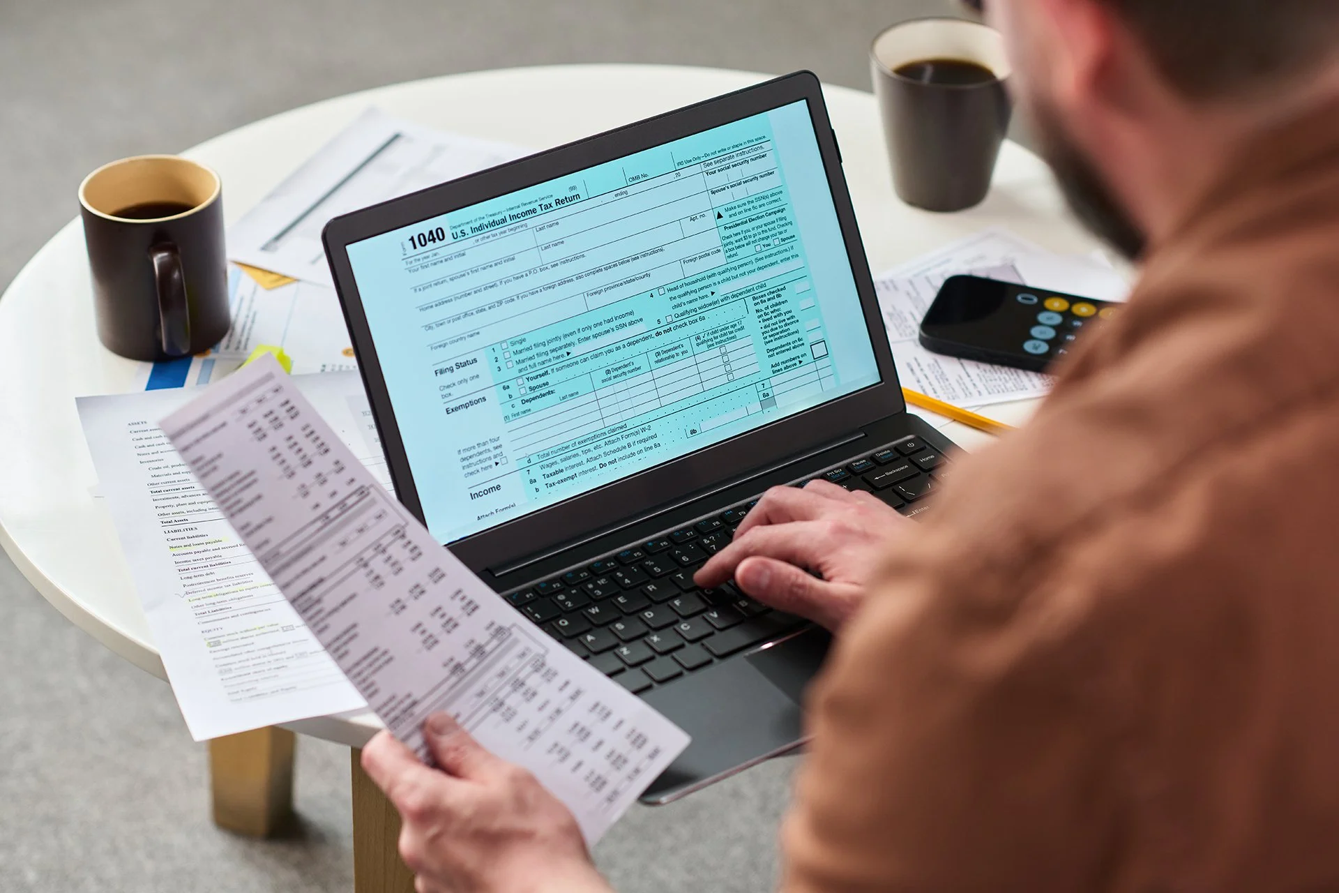 Small business owner sitting at a desk reviewing paperwork and using a laptop to file their own taxes, surrounded by receipts and financial documents.